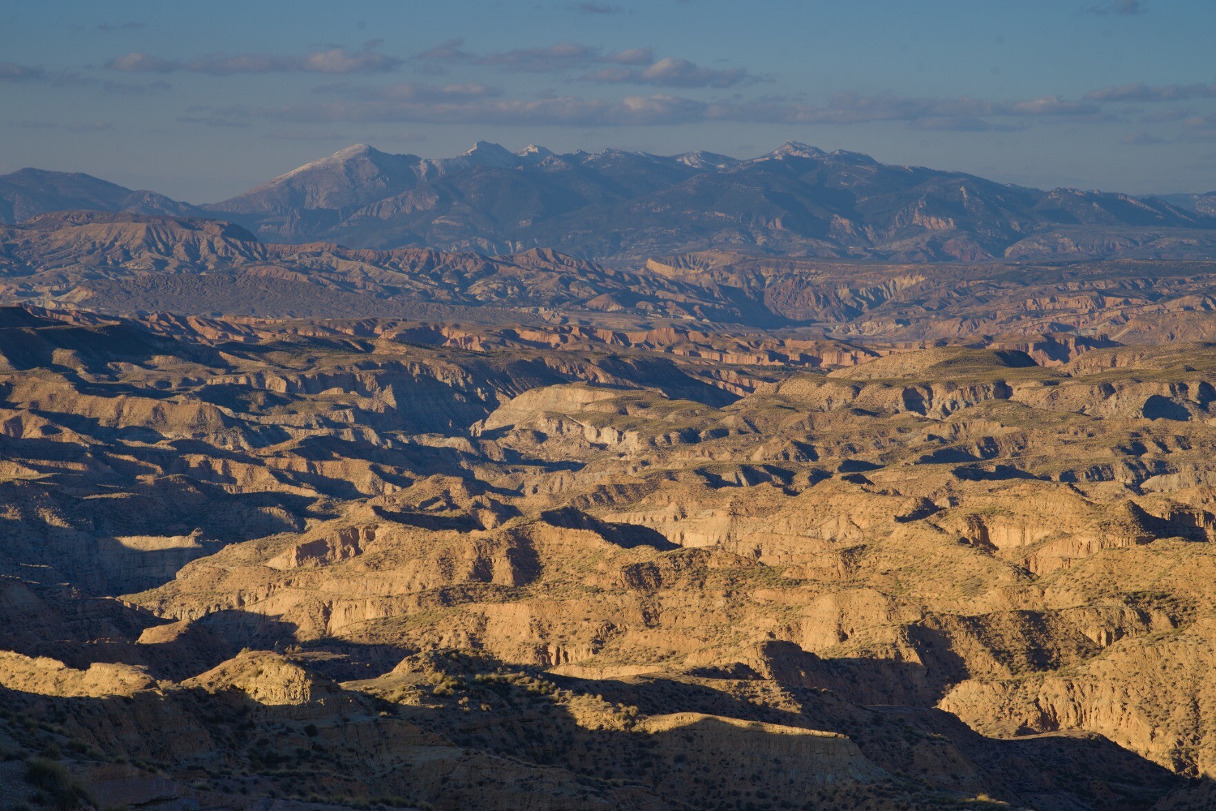 An arid desert badland landscape with hundreds of valleys. In the background some high snow mountain peaks