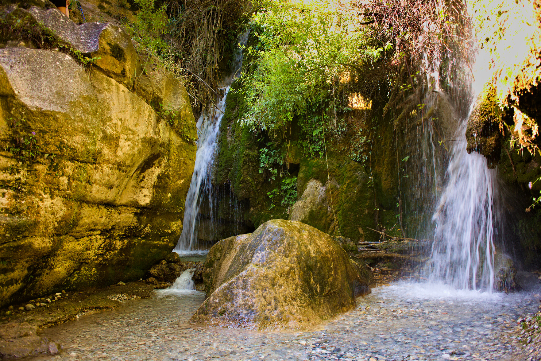 Two waterfalls plunge down from green vegetation at the top of the image. A large rock is on the left. Below a small lake forms