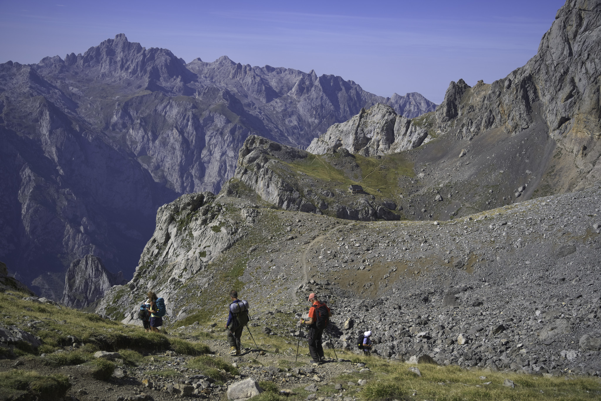 In the distance lies a grassy alpine meadow with a mountain refuge situated on it. In the foreground some walkers descend towards it.