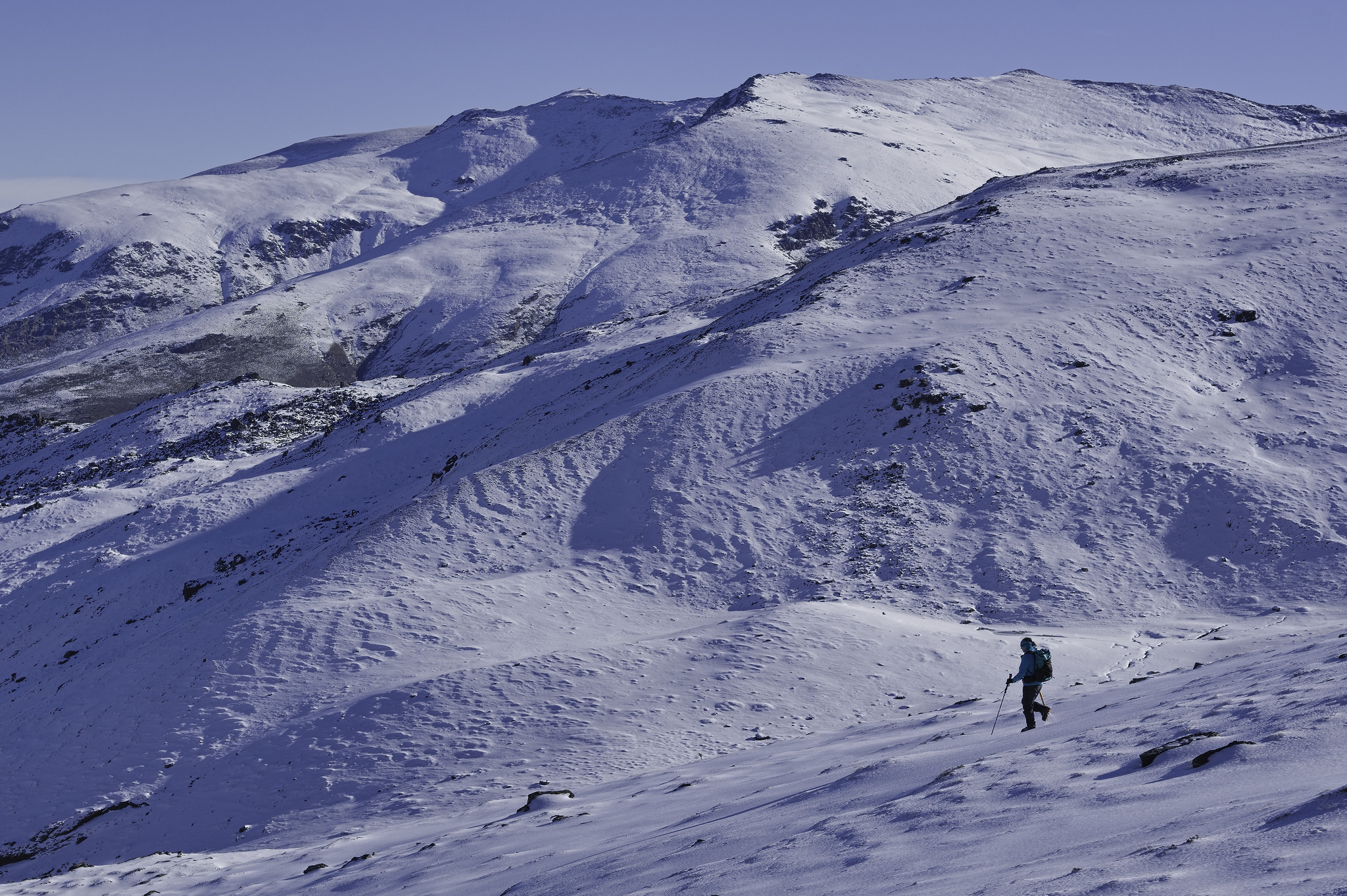 A person descends a snow slope from right to left. Behind are snow covered mountains