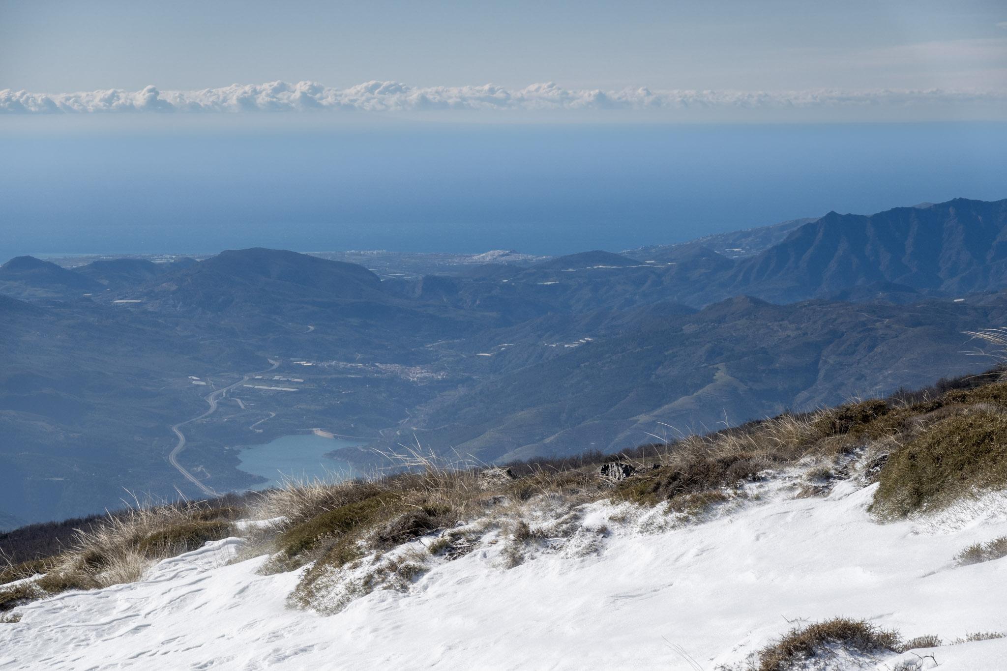 Snows in the foreground lead to grass slopes and a reservoir and a white coastline (town of Salobreña). Clouds over the sea