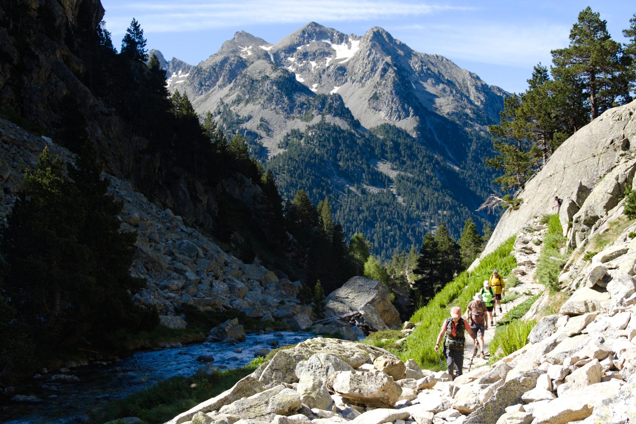 Hikers move along a sunlit track on the right. The left side of the river remains in shadow. Being rises a mountain peak with snowfields