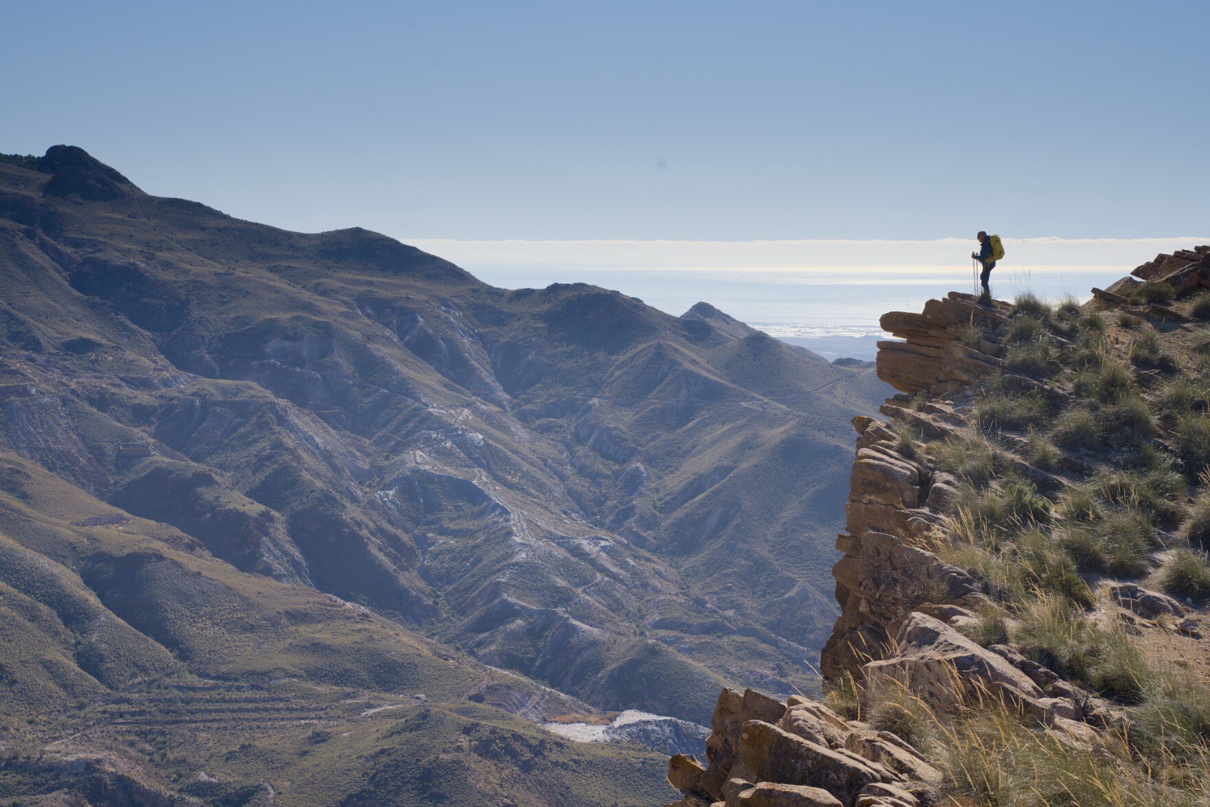 Man stood on a high Spanish mountain pinnacle with mountains and sea behind 