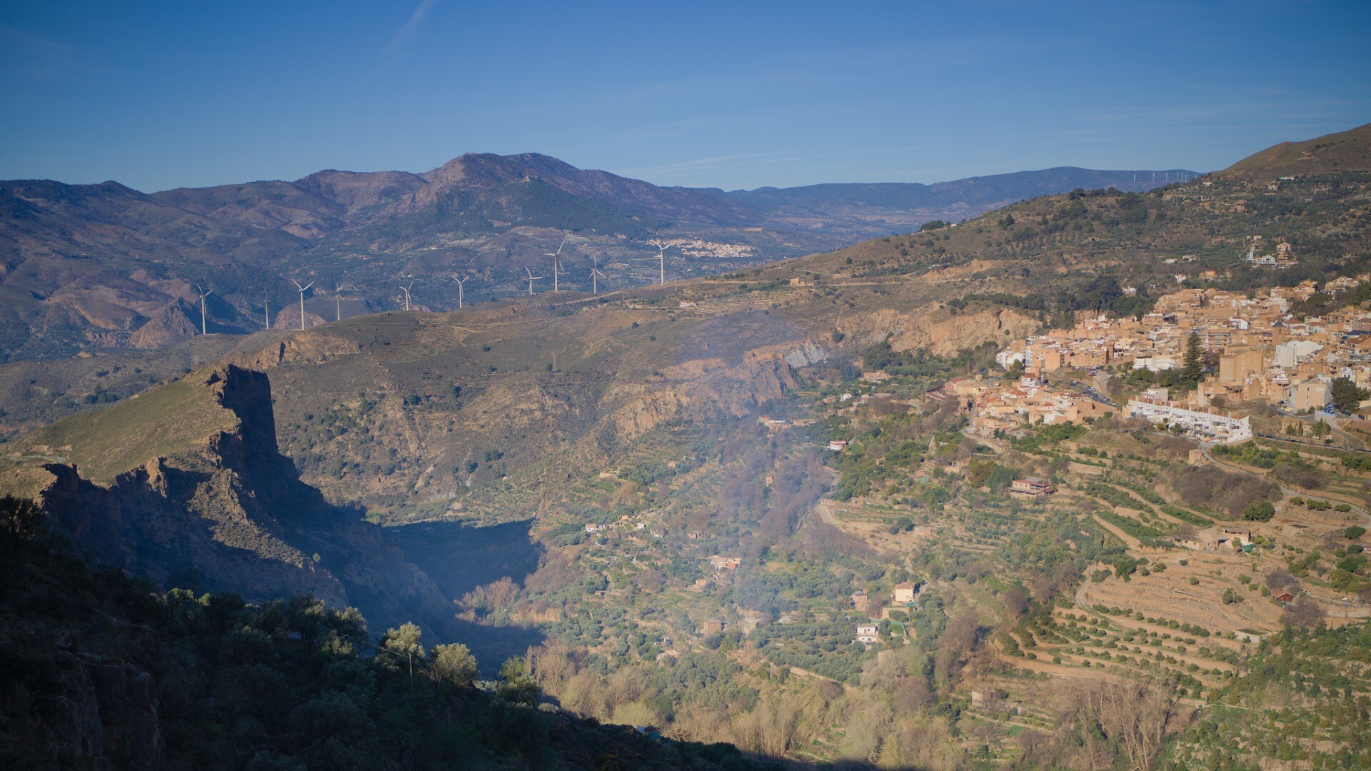To the right the spanish town of Lanjaron perched on the hillside. To the left are some shadowed cliffs. Behind a bright blue morning sky
