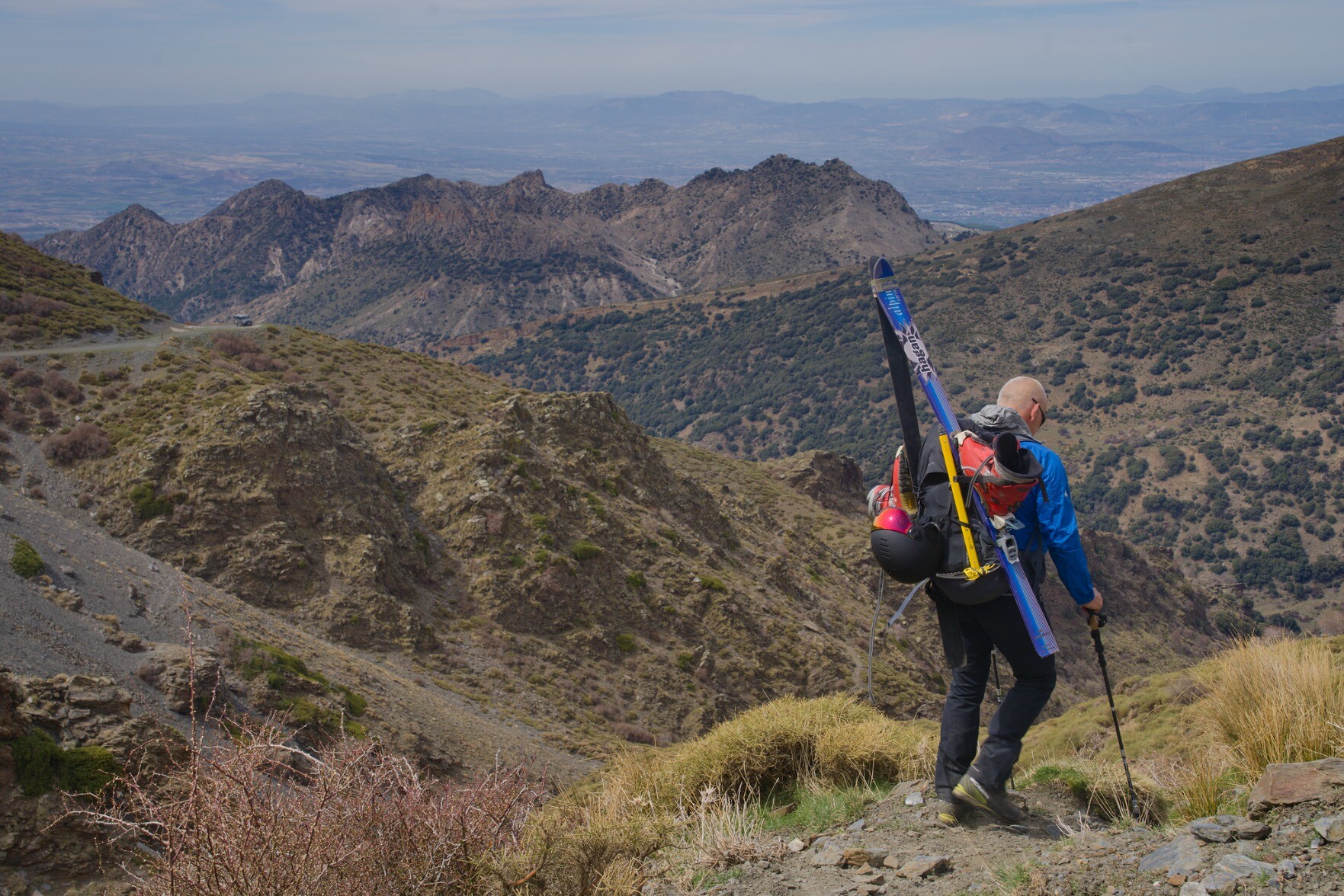 Man with skies walks down a snow free mountainside with distant views of snow free mountains
