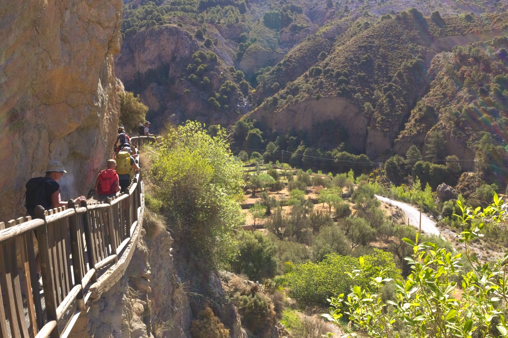 A group of hikers walk along a narrow path with a fence guarding the drop. A cliff looms high above. There is a lush green valley to the right