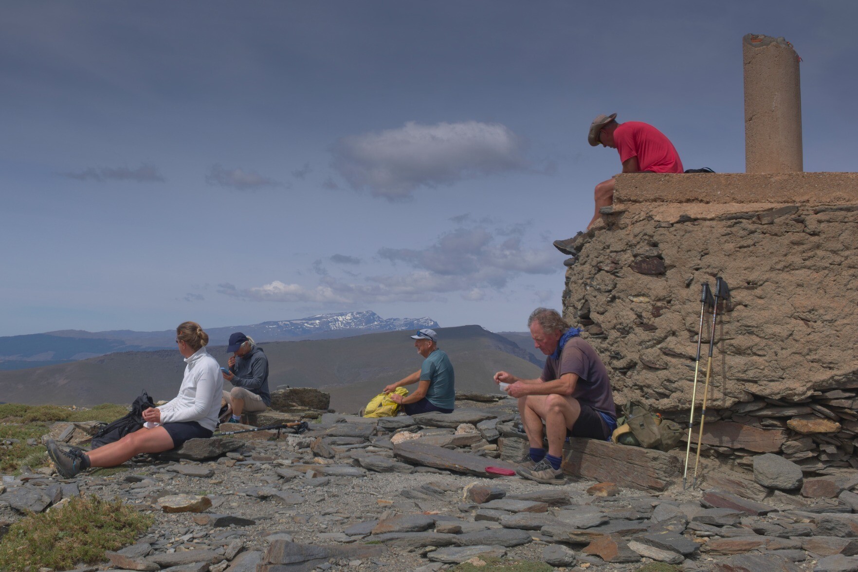 People sit and eat lunch on a mountain summit. There is a large trig point to the right. In the distance are some snow covered peaks