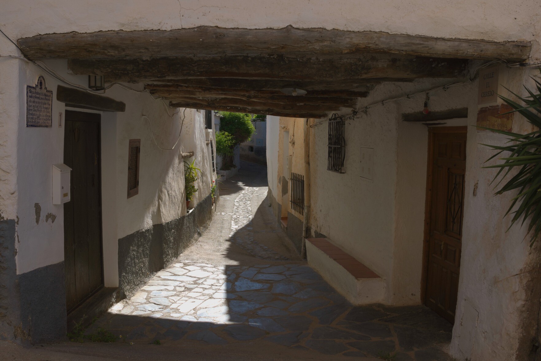 A narrow alleyway with old buildings on both sides leads to a sunlit exit of the street with green shrubs