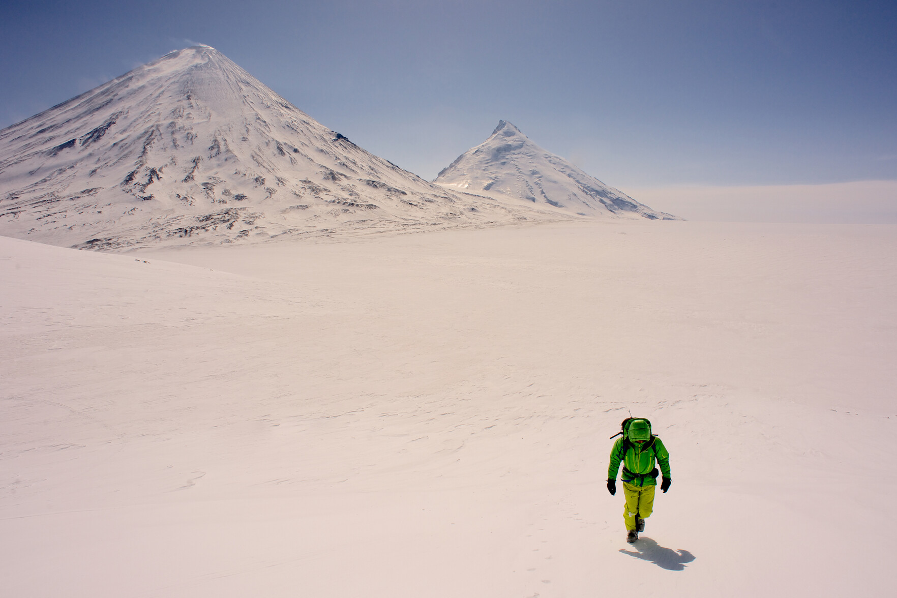 In the foreground is a man dressed in green and yellow that stands out from the surrounding snow. At the back rise two snow clad, classic shaped, mountains. Blue skies