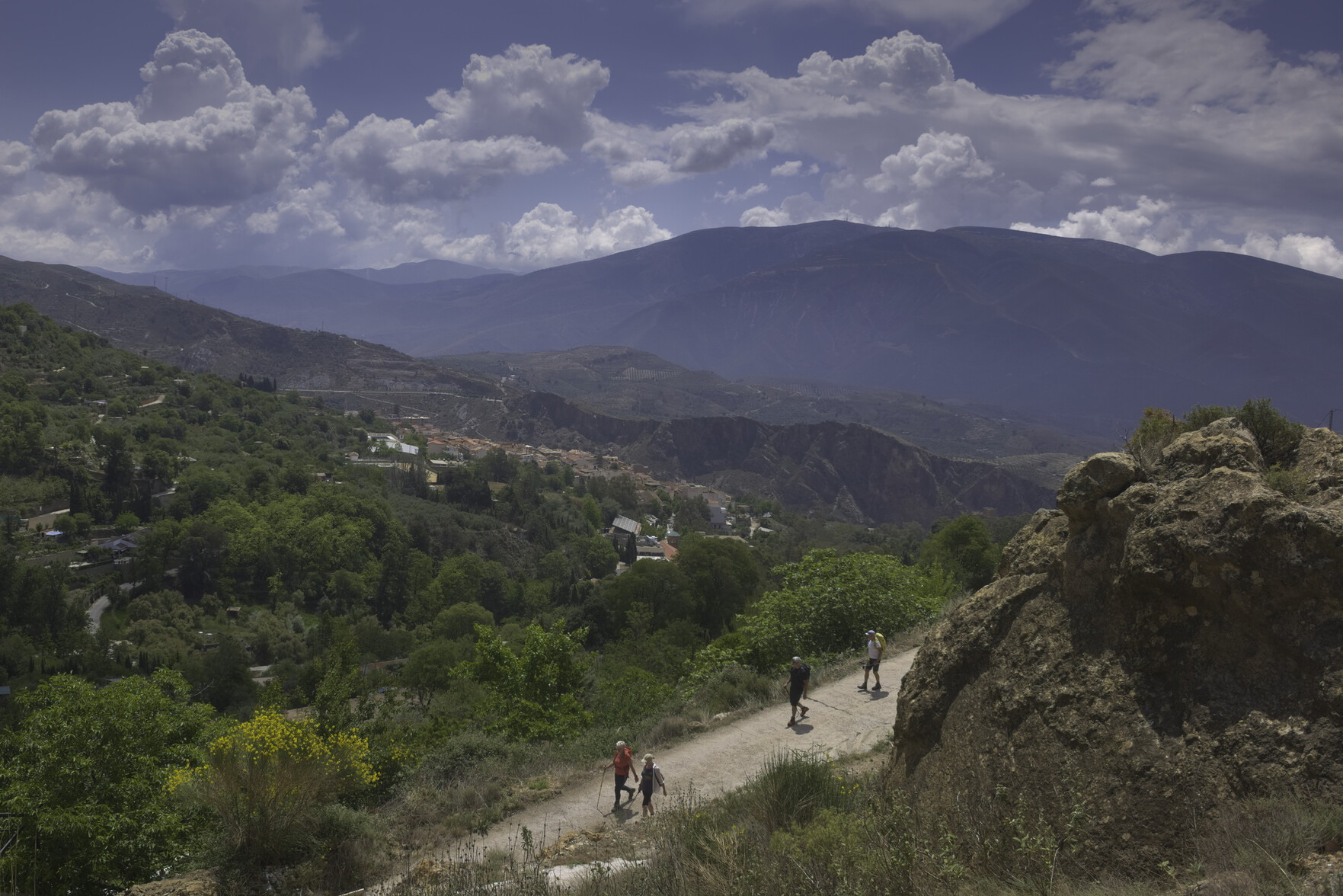 Some hikers on a dirt track in the foreground. In the middle distance  lies a town. Behind are a range of mountains with clouds above them