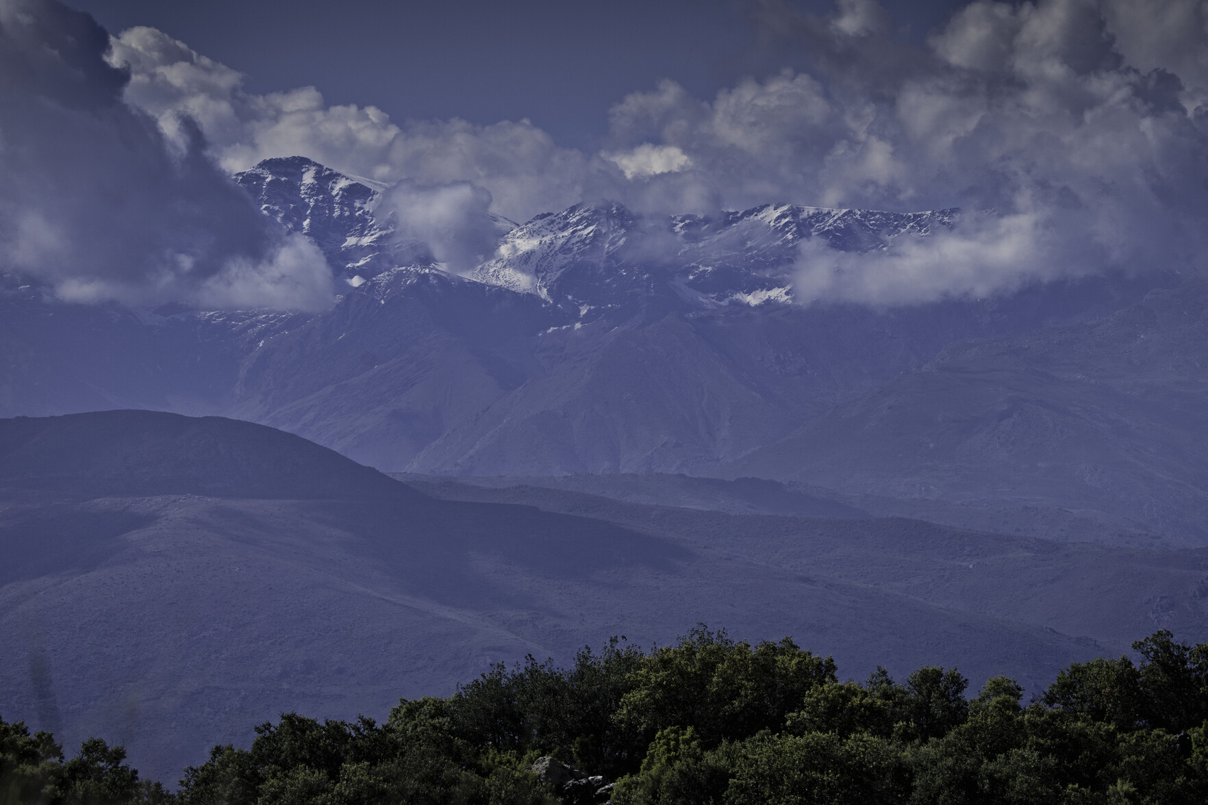 In the foreground some green tree tops. behind and some high snowy mountains with cloud around them