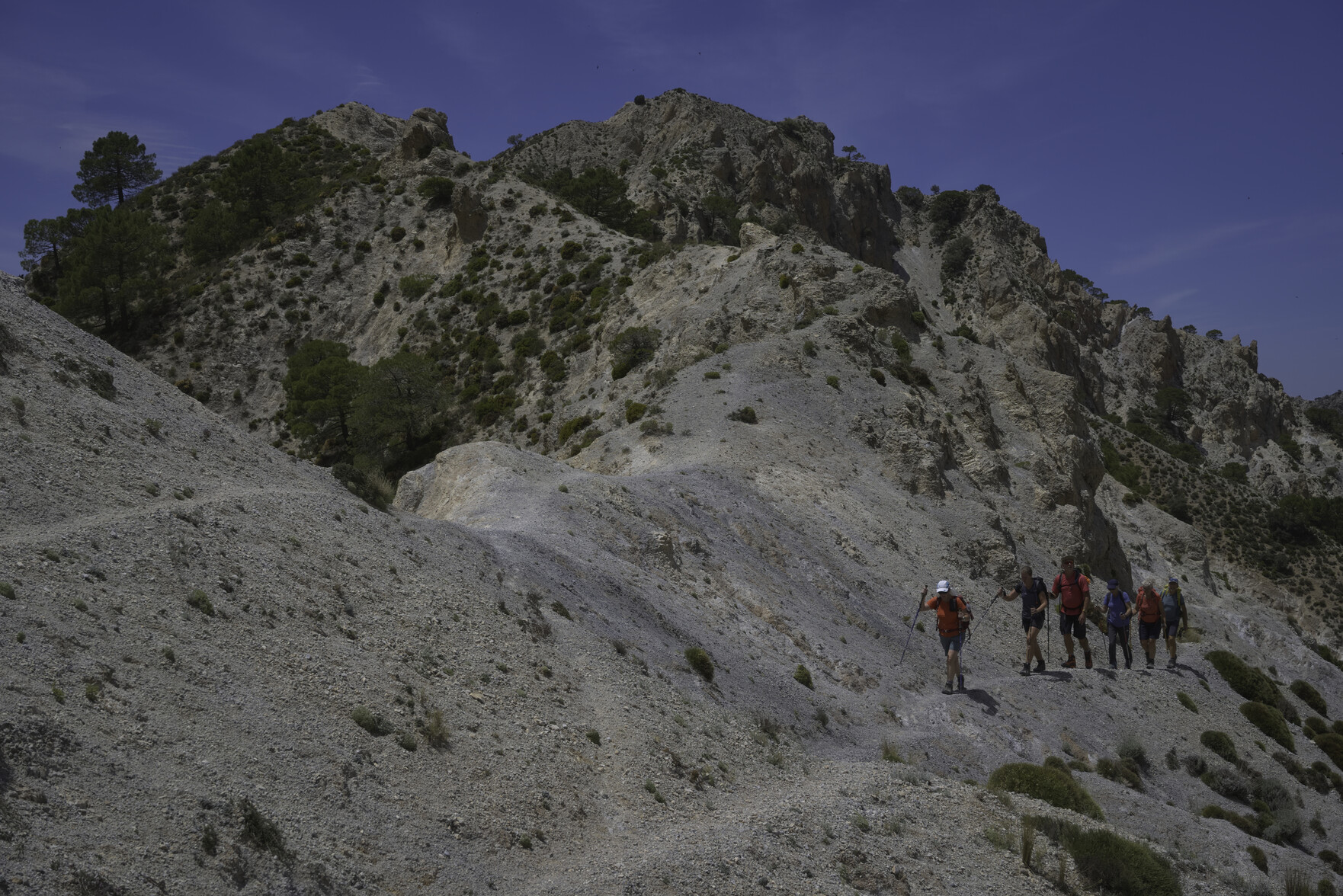 A group of hikers pass along a narrow sandy path. Up and to the left are some rocky mountain peaks