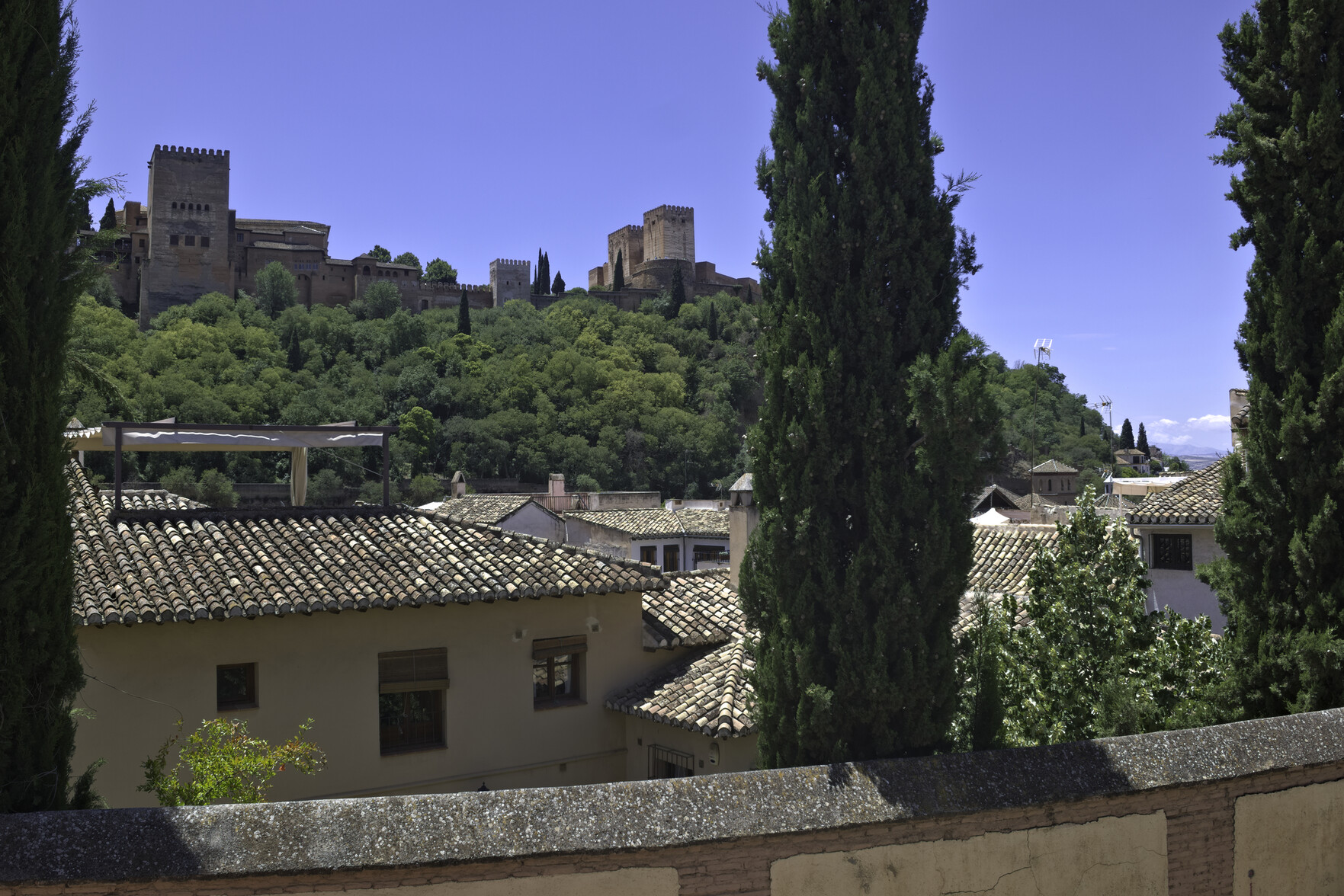 The turrets of the Alhambra Palace fortress area rise up into the blue sky. The palace is surrounded by green forests. In the foreground are some houses.