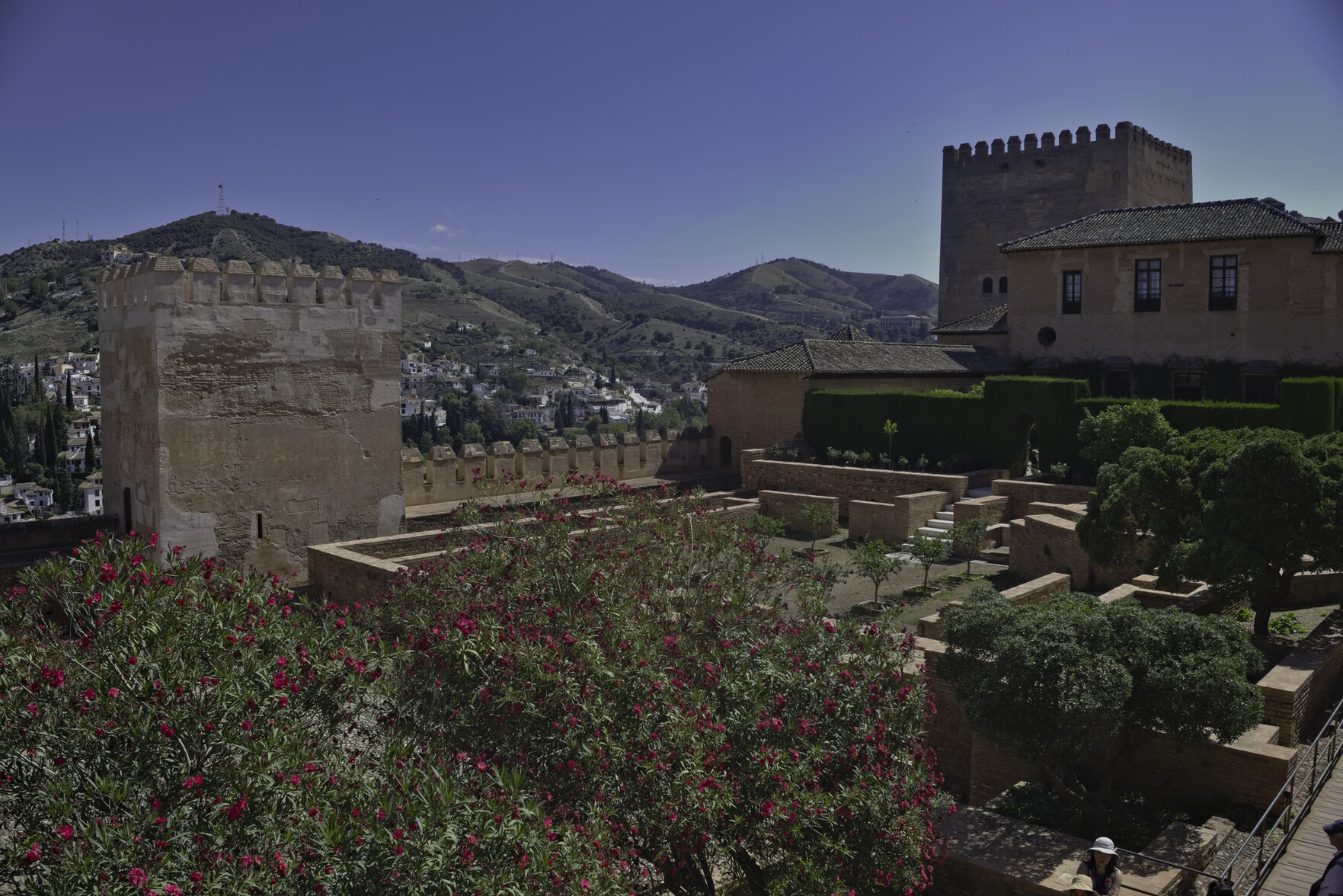 The "Torre de Muhammad" and gardens lead to the Nastrid Palaces on the right. In the forground some colorful plants. behind the building is the Sacramonte area of Granada