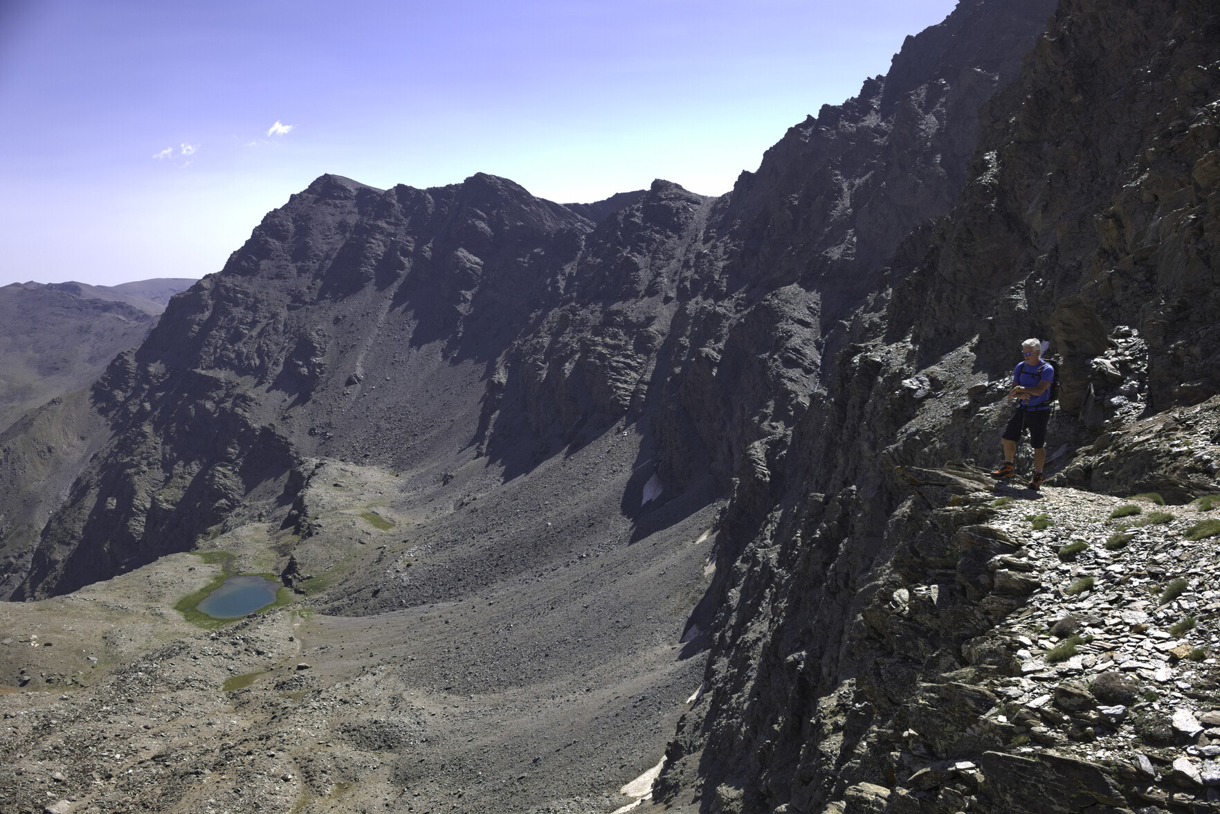 A man in a blue shirt stands perched above an enormous drop to the left down to a small blue lake