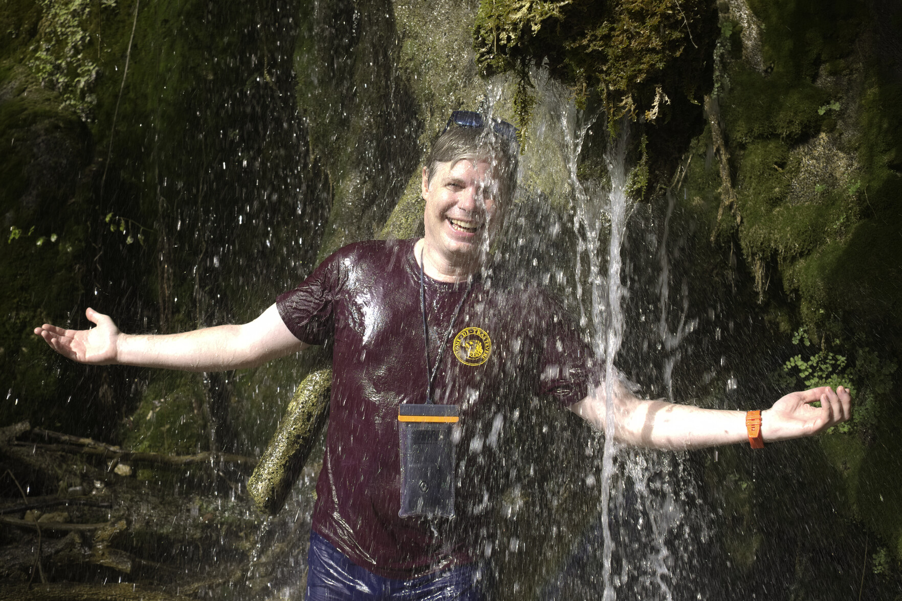 A man in purple tee shirt stands under a waterfall