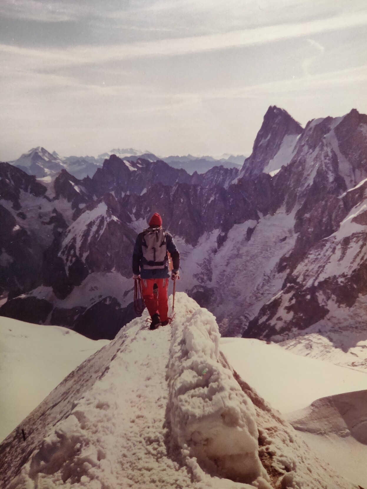 A man with red pants is seen on a narrow snow ridge in the foreground. Behind are sharp, dramatic looking dark peaks