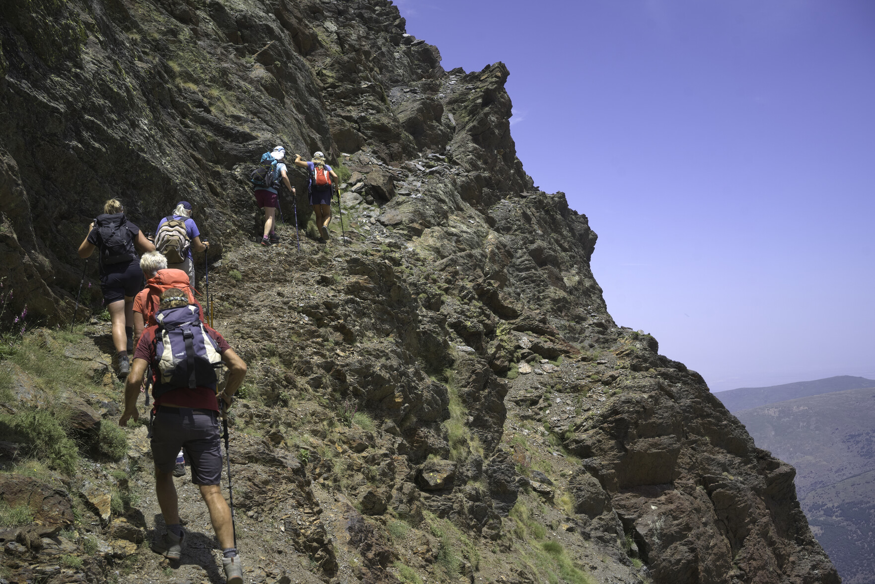 A group of hikers on the left pass along a narrow path that traverses up across the face of a steep mountainside covered with cliffs