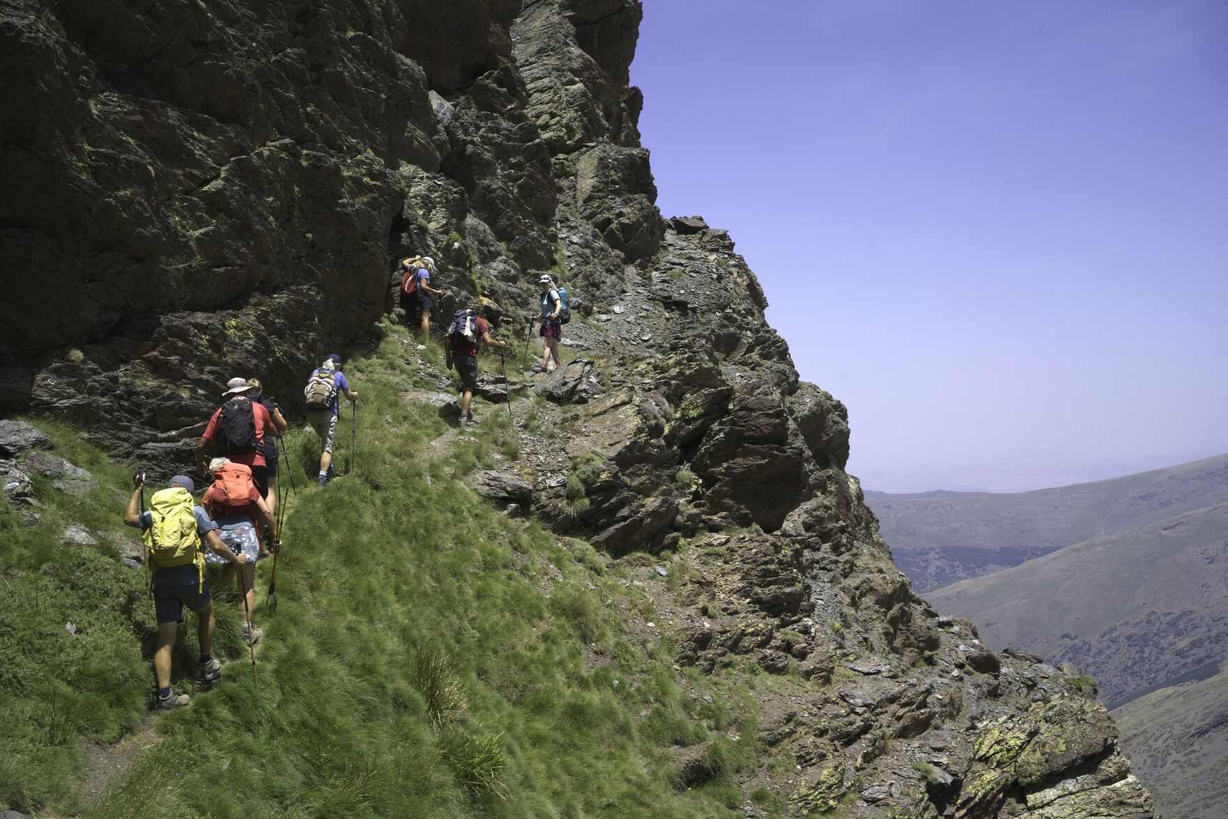 A group of hikers on the left pass along a narrow path that traverses up across the face of a steep mountainside covered with cliffs