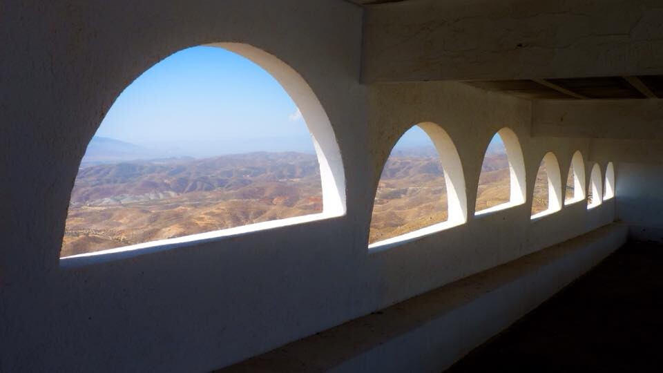 A building has semi circle windows extending down the left hand edge. Through the windows can be seen images of desert badlands and blue sky