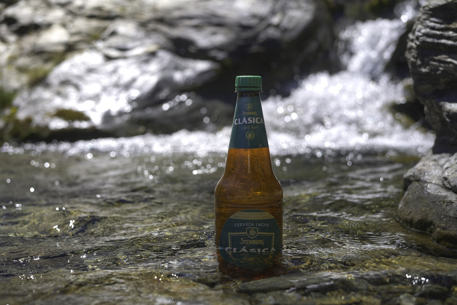 A bottle of beer sits cooling down in a mountain stream. A small waterfall is to the right