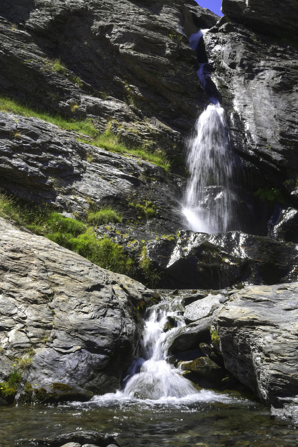 Water flows down over a waterfall on the right into a pool at it's base. Some green grasses adorn the sides