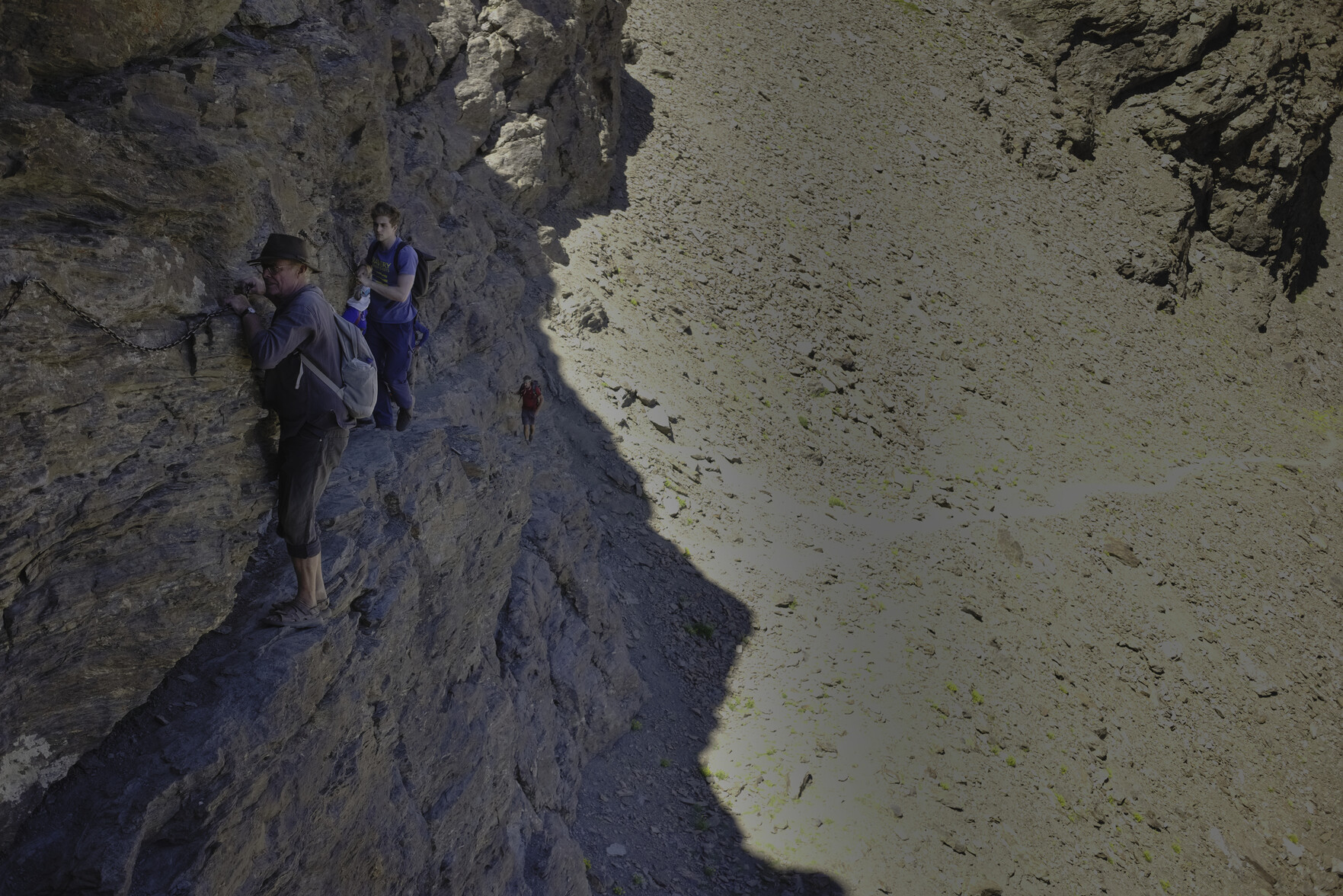 A man on the left moves slowly across a very narrow rocky ledge whilst holding onto a chain fixed to the rock wall. To the right the rock  falls away vertically to the valley floor