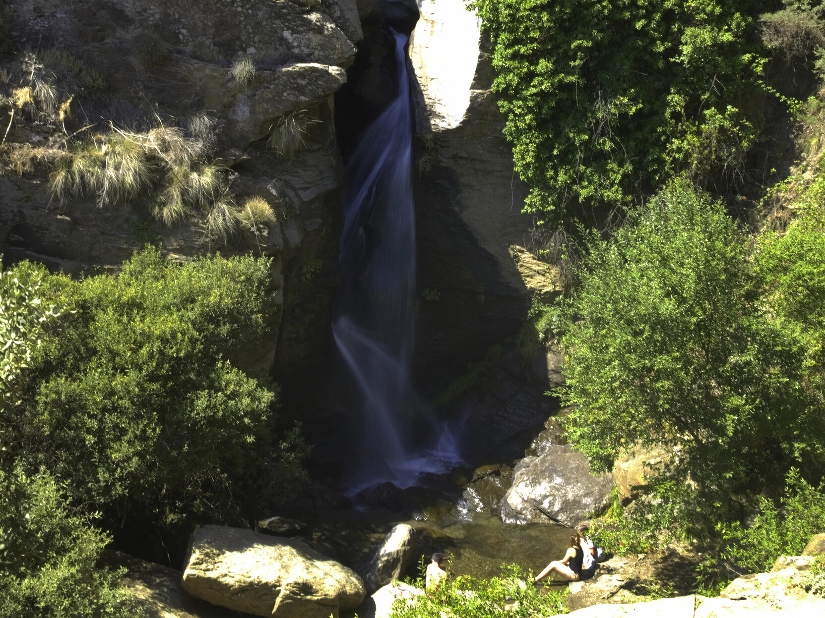 In the center of the image, water tumbles down a narrow cleft in the rocks. To the sides there is lush green vegetation. Some people sit at the base of the waterfalls