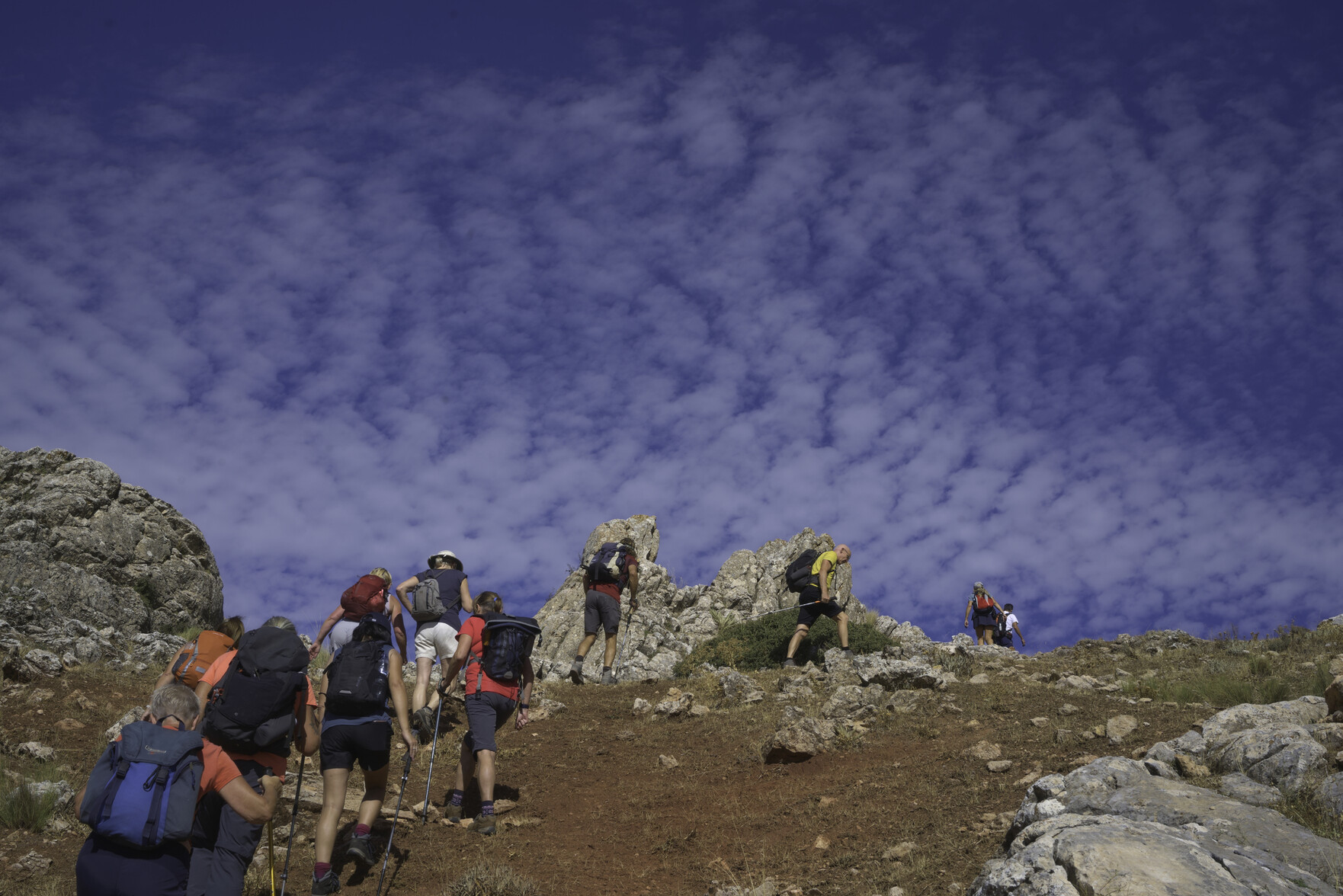 A group of multi coloured hikers walks beneath a beautiful blue sky covered with white mackerel clouds