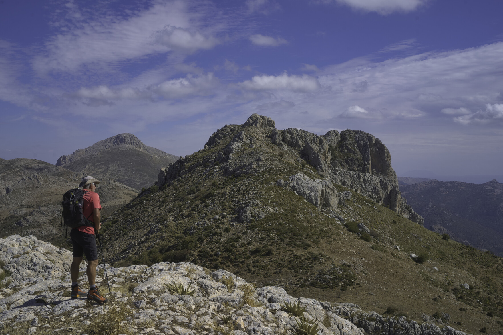 A lone hiker looks towards some distant mountains. Above a blue sky with some white clouds