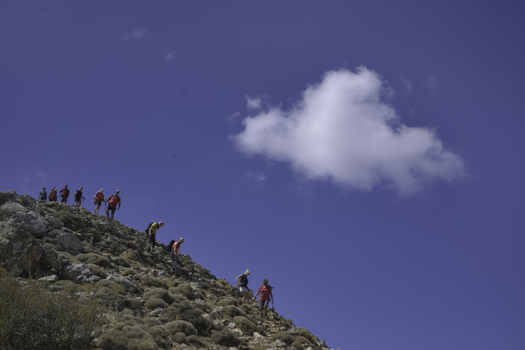 Hikers descend steep ground to the left. Above a blue sky with a solitary white cloud 