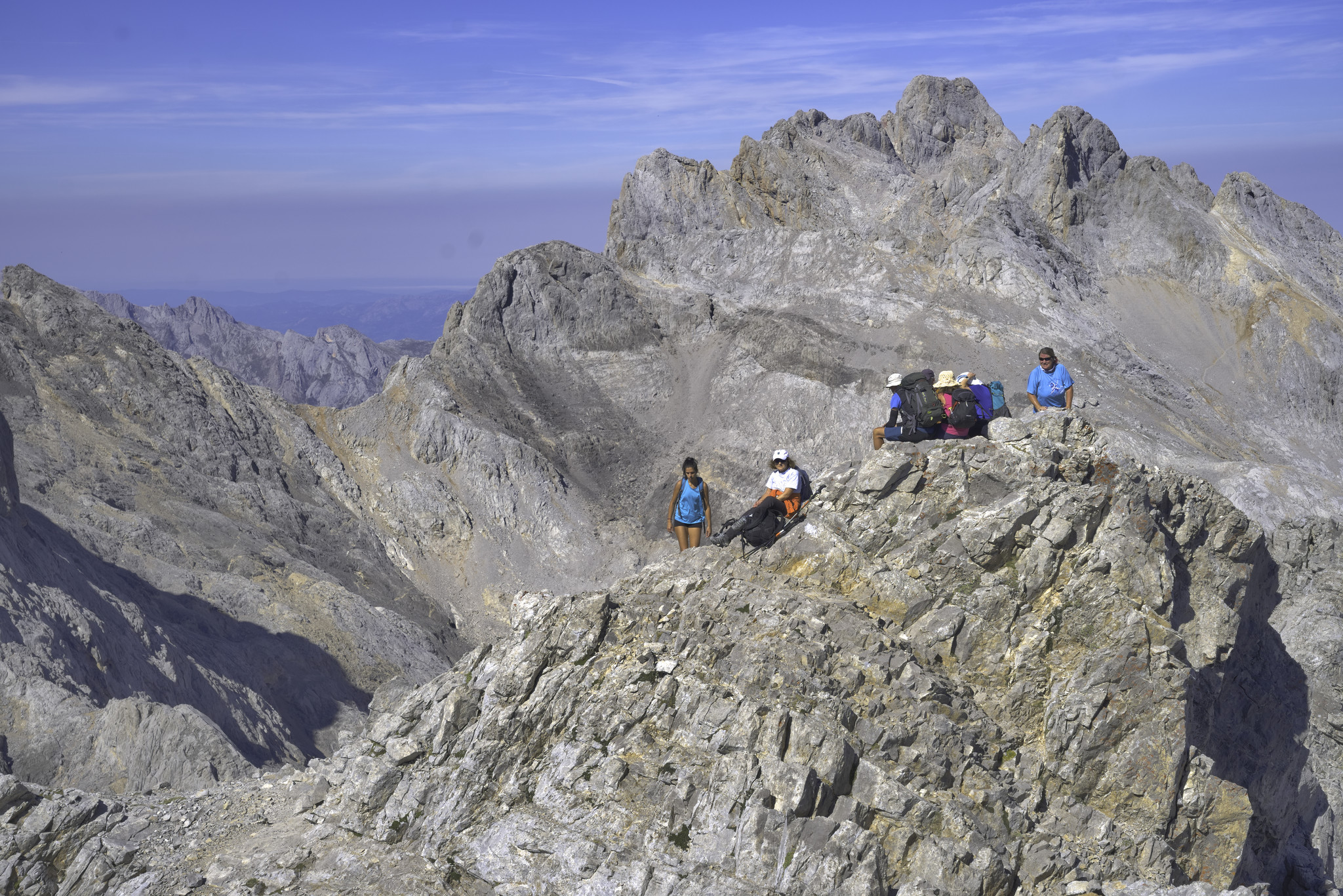 A colorful group sit on the narrow summit of Torre de Horcados Rojos. Behind rise other mountains with blue sky above
