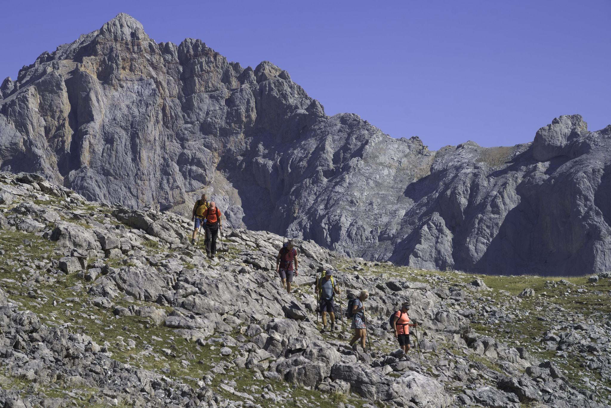 A group of hikers descend some rocky ground, behind and to the left rises a huge mountain wall