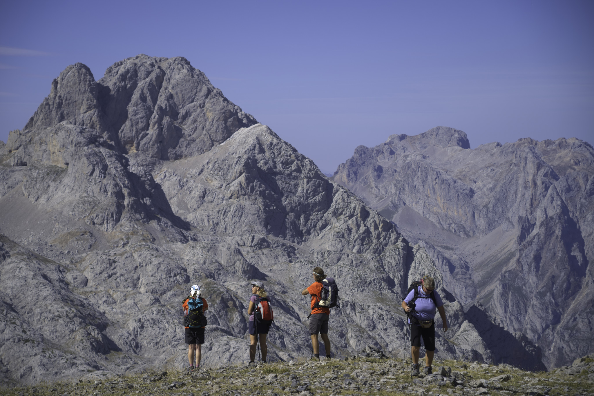 Three walkers in red stand looking at a huge mountain rising behind them. Blue sky all around