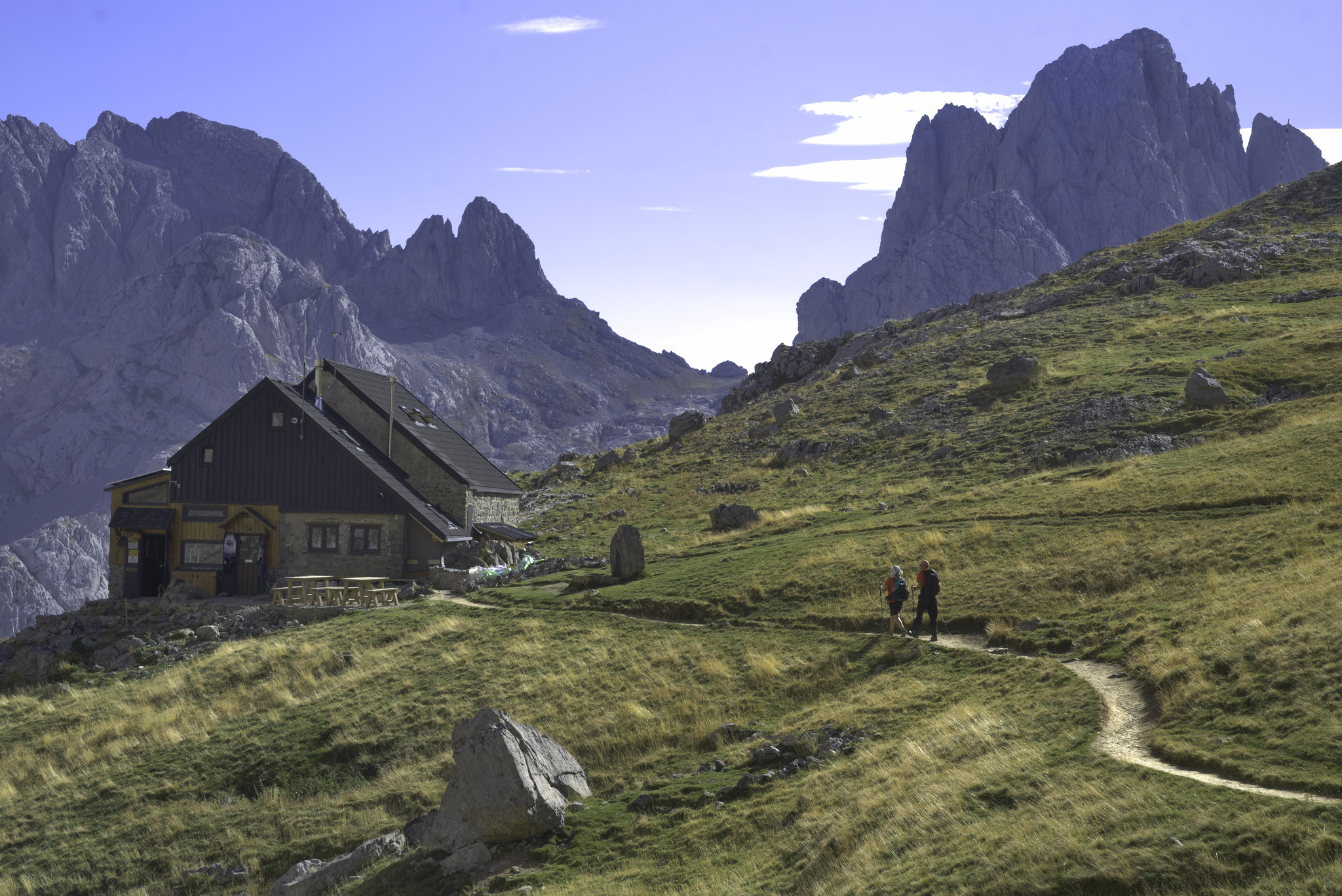 A mountain refuge sits amid a grassy alpine meadow with two hikers walking along a path to it. behind rise high dramatic mountains