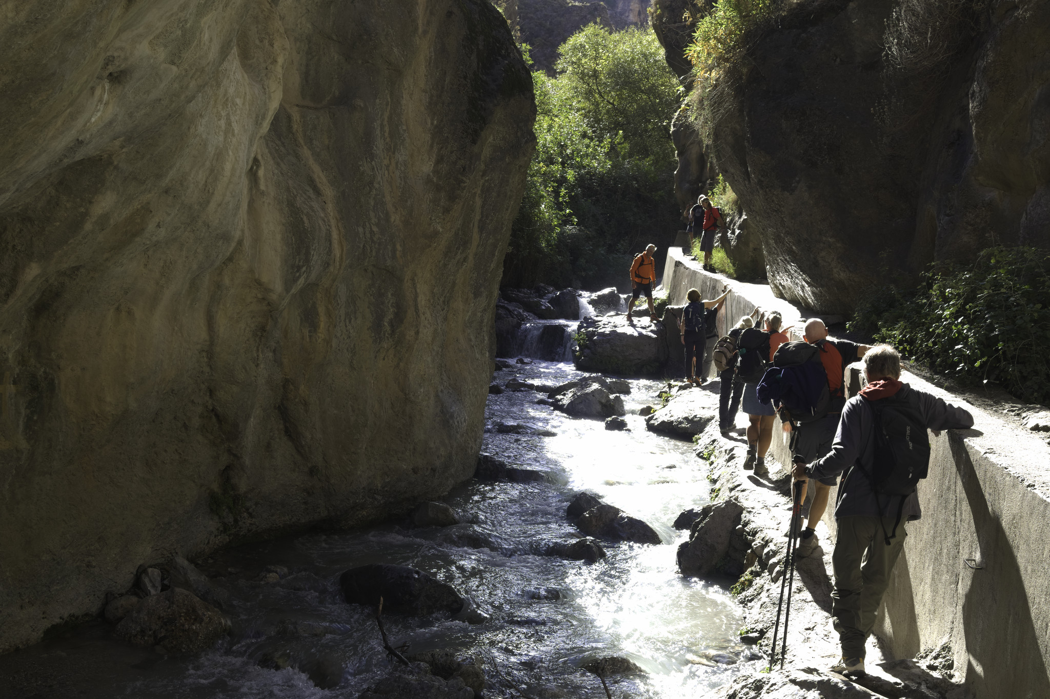 Some people on the right pass along a narrow path next to a river. A large ochre colored rock wall is to the left of the river