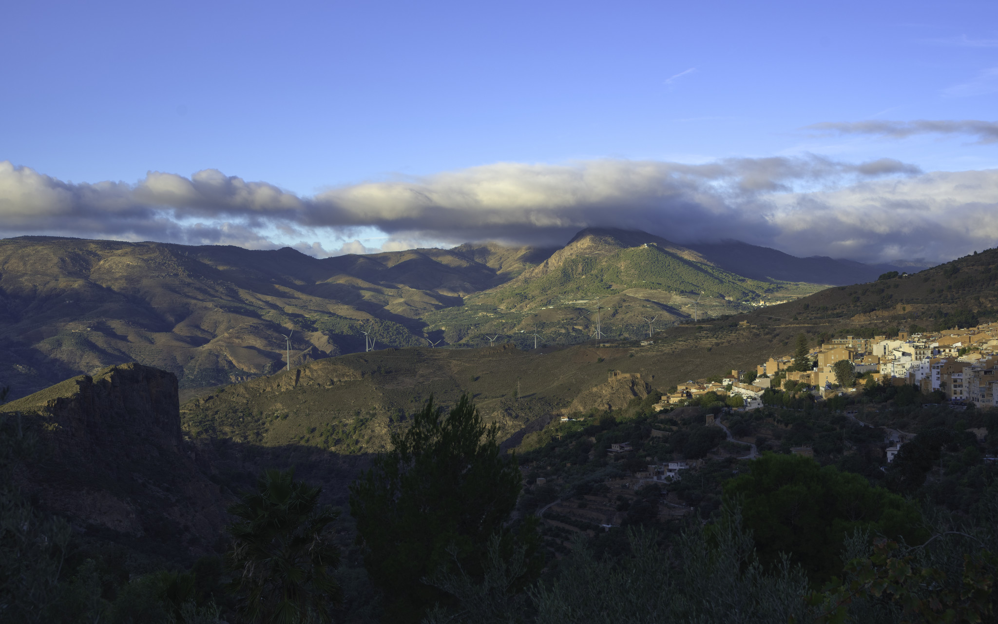 A small spanish town on the right gets the first rays of autumnal sunshine. Behind lie green and brown hills with clouds and blue sky above