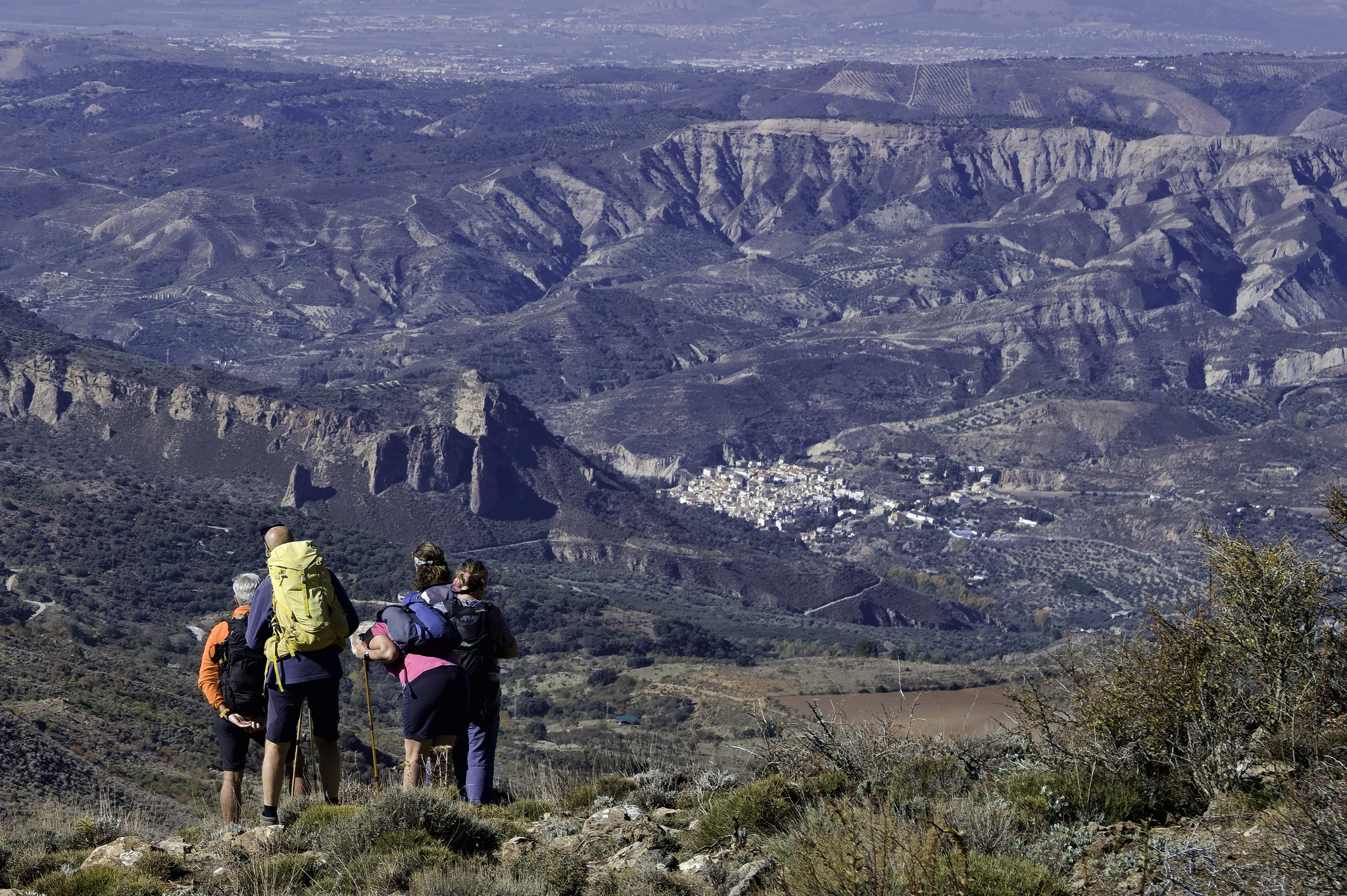 A group of hikers are at the bottom left. Behind them are arid craggy slopes and the small town of Quentar