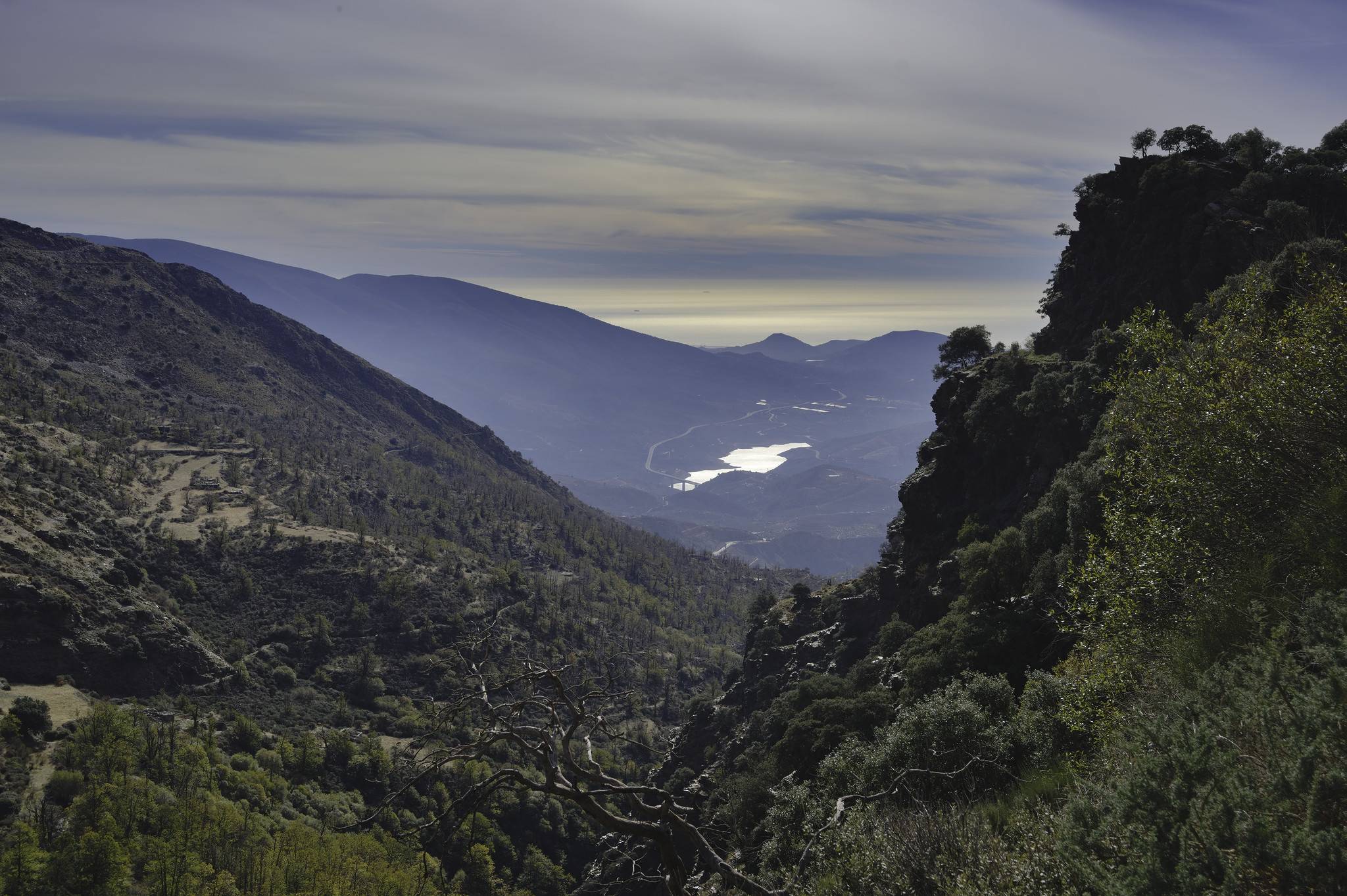 A long distance view to the Mediterranean coast. A V shaped valley with a prominent craggy outcrop on the right. Wispy clouds over the sea
