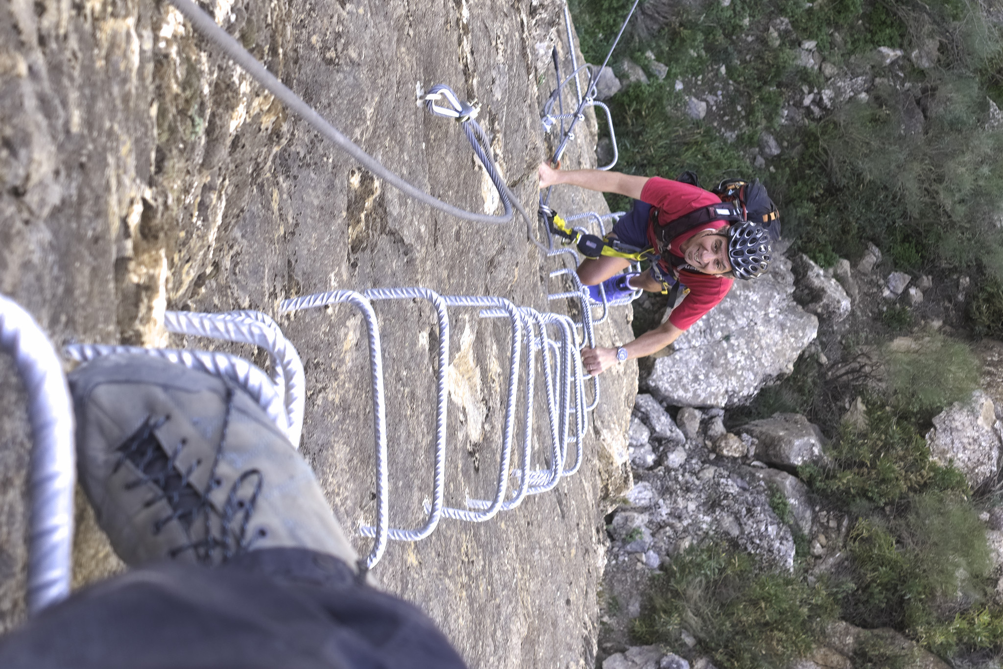 A person is climbing up or rock wall equipped with ladders and using cables for protection. A persons foot (mine!) is at the bottom left of the photo standing on a ladder rung