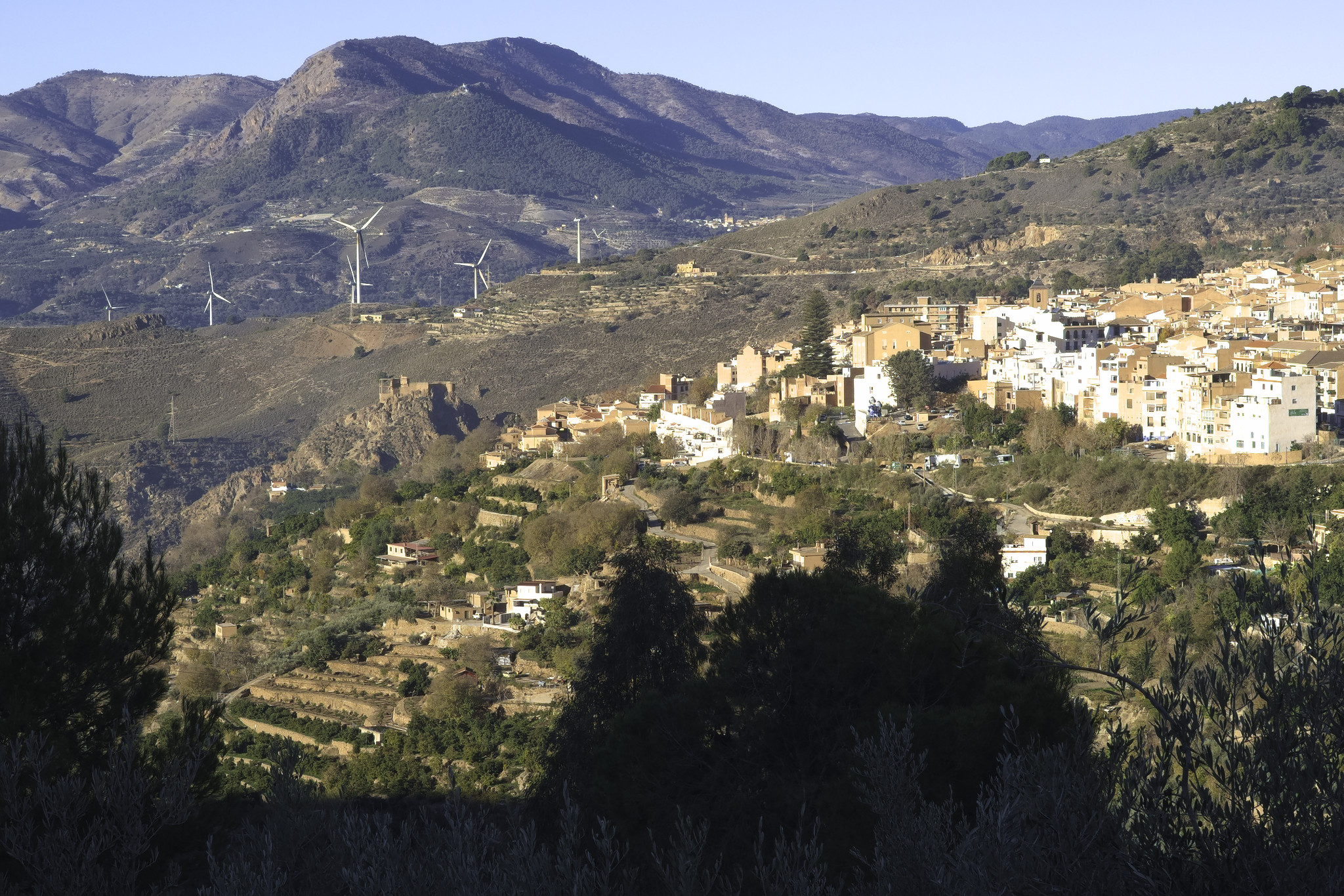 A town sits on a sunlit shelf to the right. To the left of the town is a ancient arab castle. behind are some wind turbines and a range of mountains.