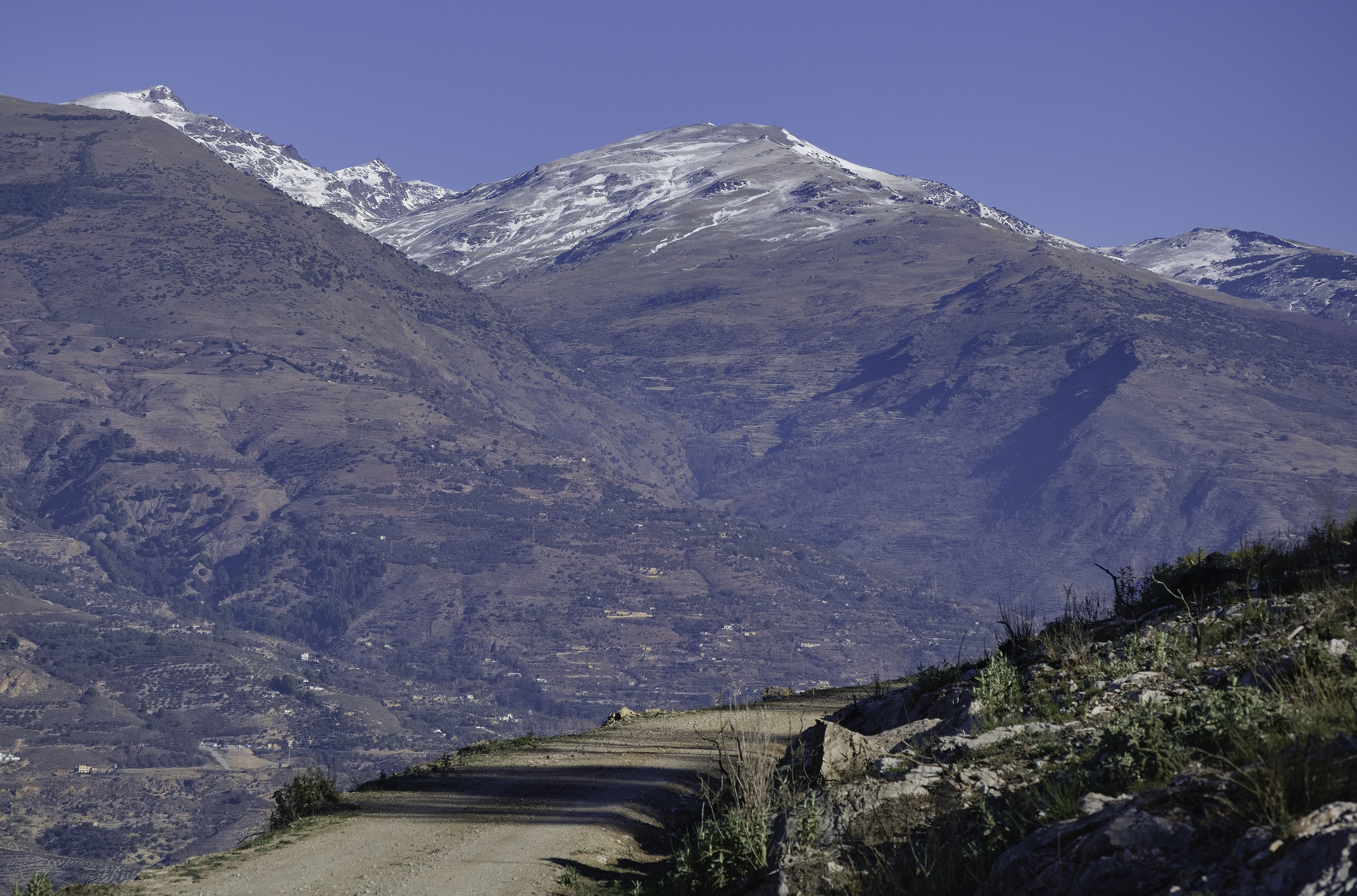 A dirt road in the foreground leads the eye to the mountains behind which become snow covered towards the summits