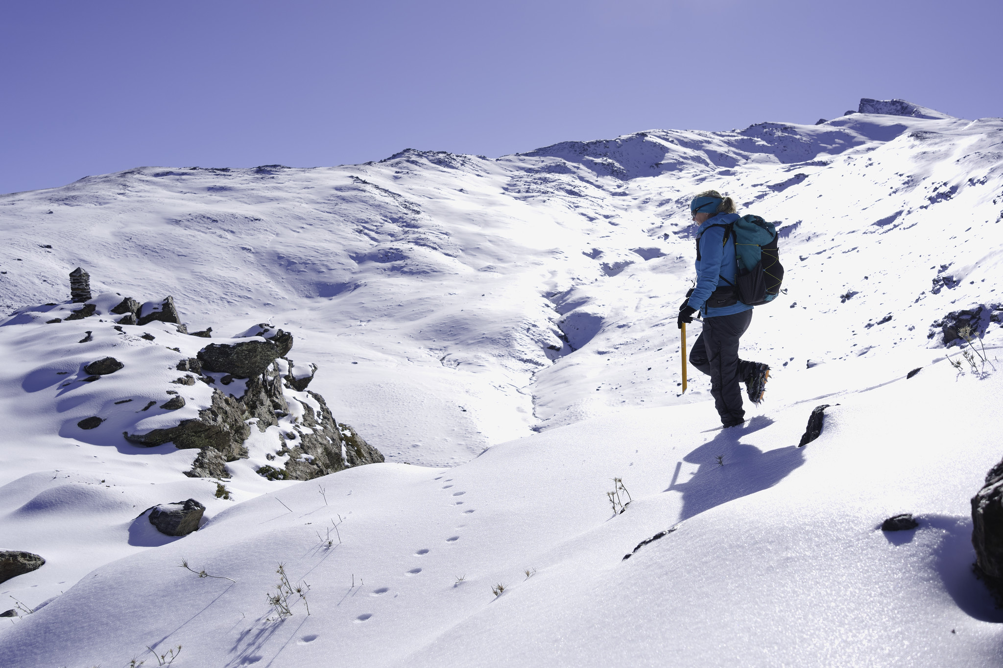 A person dressed in a pale blue jacket descend a slope from the right towards a small summit with a large cairn on. Behind rises a big mountain