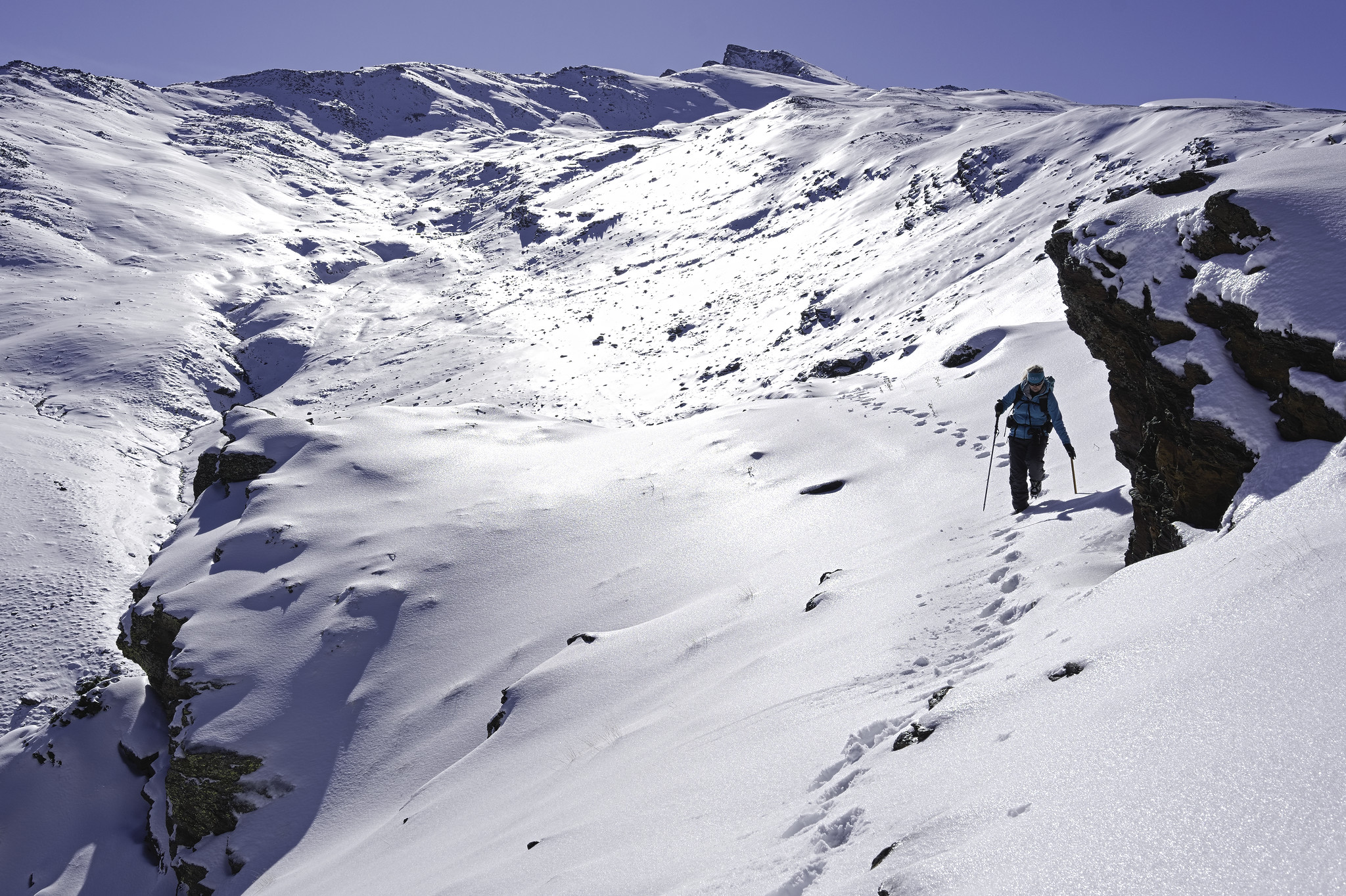 A person follows footprints in the snow to the right of the image. The person is beneath a large boulder covered in snow. To the left the cliff drops off vertically