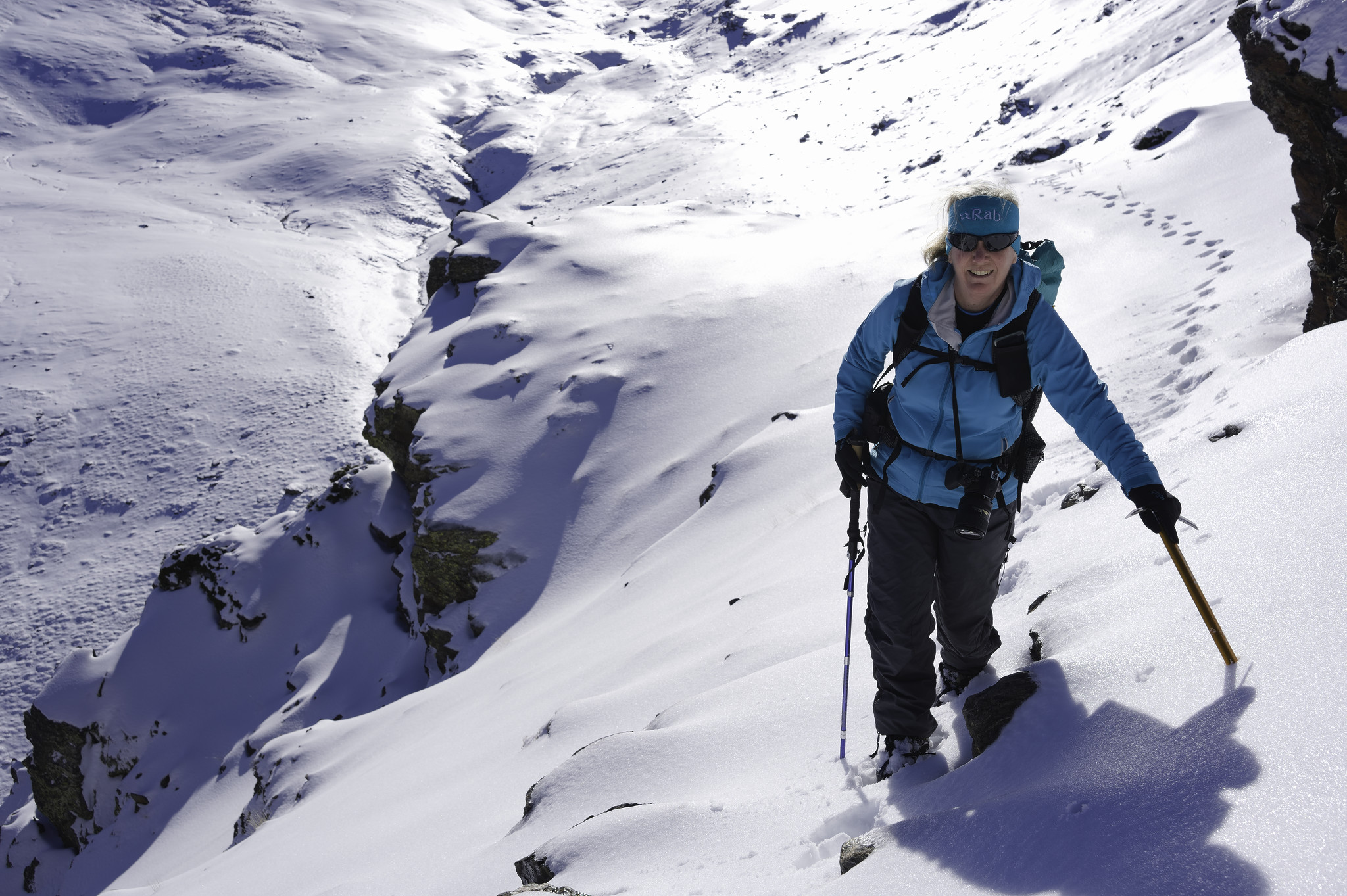 A person in a sky blue jacket moves along a small snow covered path carrying an ice axe and hiking pole. To the left a cliff edge is shown