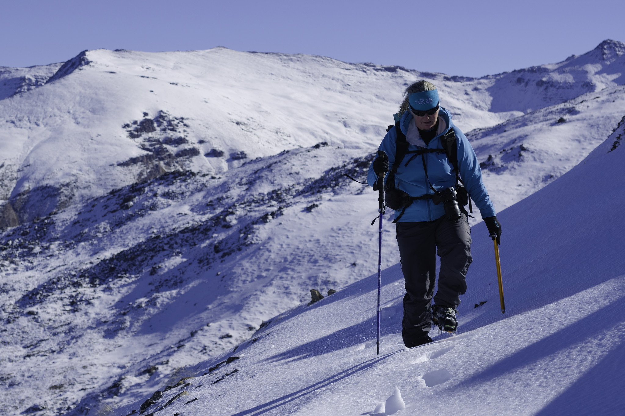 A person in a sky blue jacket moves along a small snow covered path carrying an ice axe and hiking pole. Behind are some higher snow covered mountains with blue sky above 