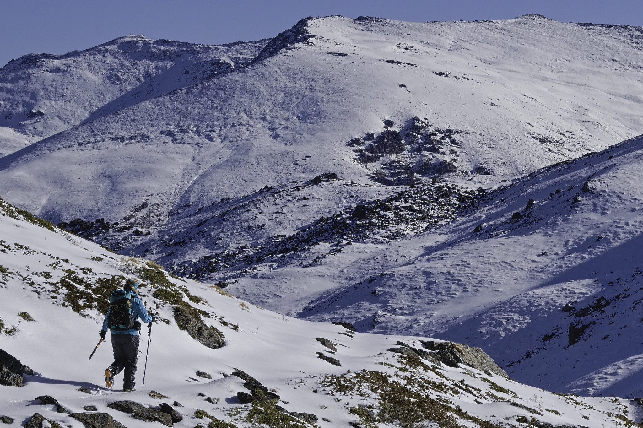 A person in a sky blue jacket moves along a small snow covered path on the left. Above and to the right is a large snowy mountain with some small forested sections.