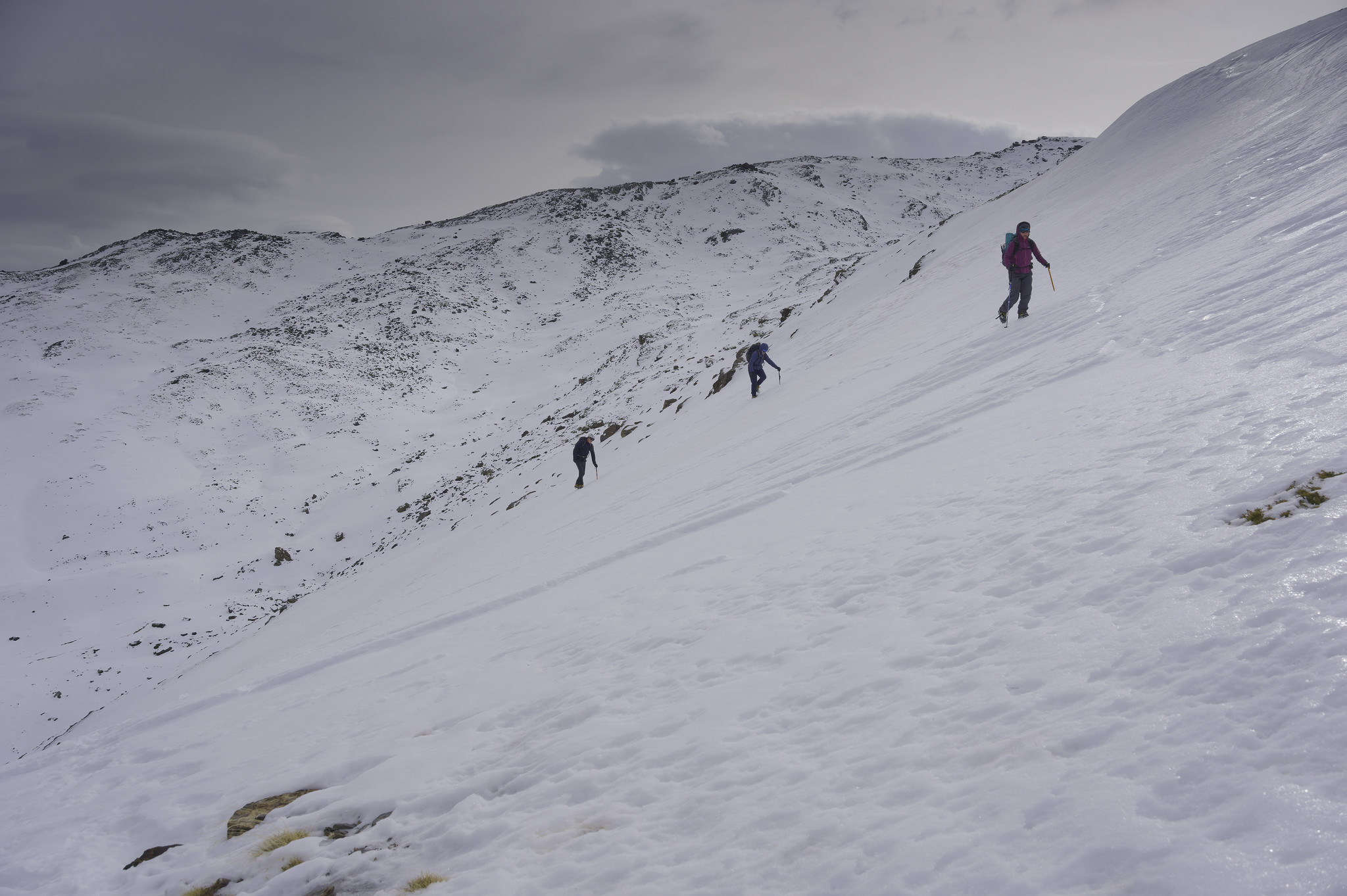 Three people are climbing up a steep snow slope on the right of the photo. Behind them rise other mountains covered in snow and ice