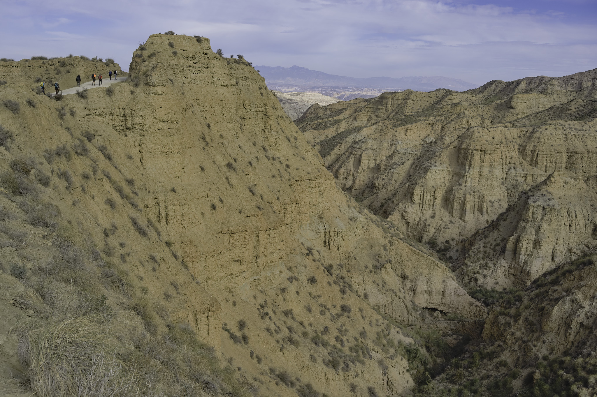 A small group of hikers walks along a wide path in the upper left of the image. Below and to the right is a  mix of orange cliffs and crags that lead into a valley. Badlands!