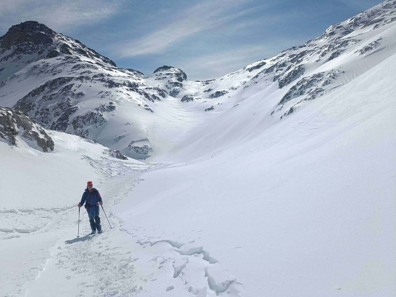 A person dressed in blue and in snowshoes is surrounded by snow and rugged high mountains. Blue sky above